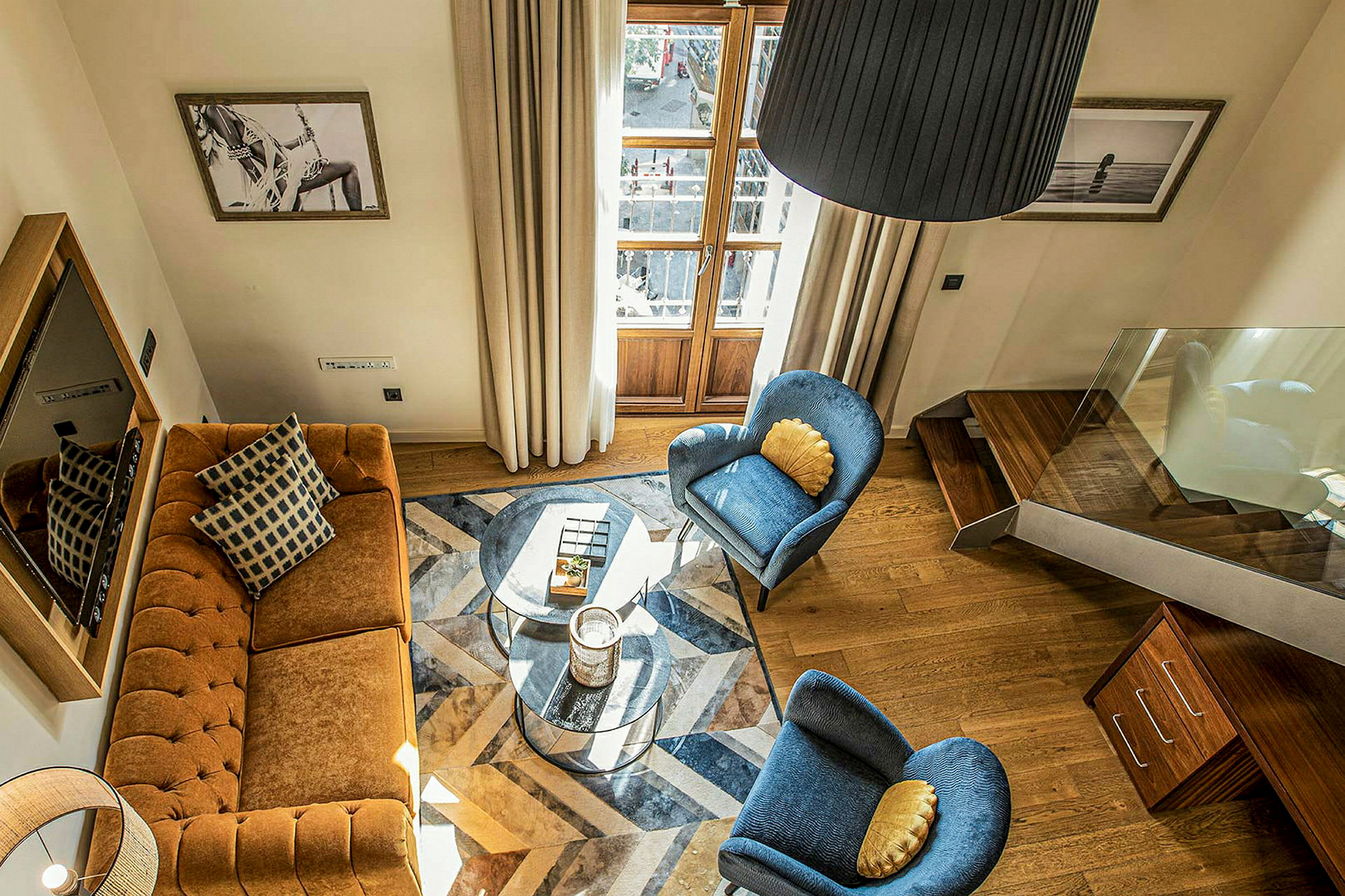 Et opholdsrum med en brun sofa, to blå stole, et glasbord og trædetaljer. Malerier hænger på væggene. (AI-genereret tekst)