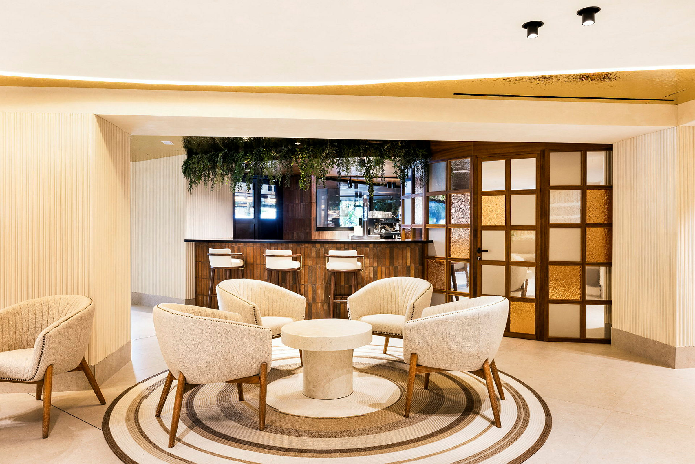 Et moderne hotelområde med en siddegruppe bestående af bløde lænestole omkring et rundt bord og en bardisk i baggrunden. Indretningen er lys og elegant med planteudsmykning. (AI-genereret tekst)