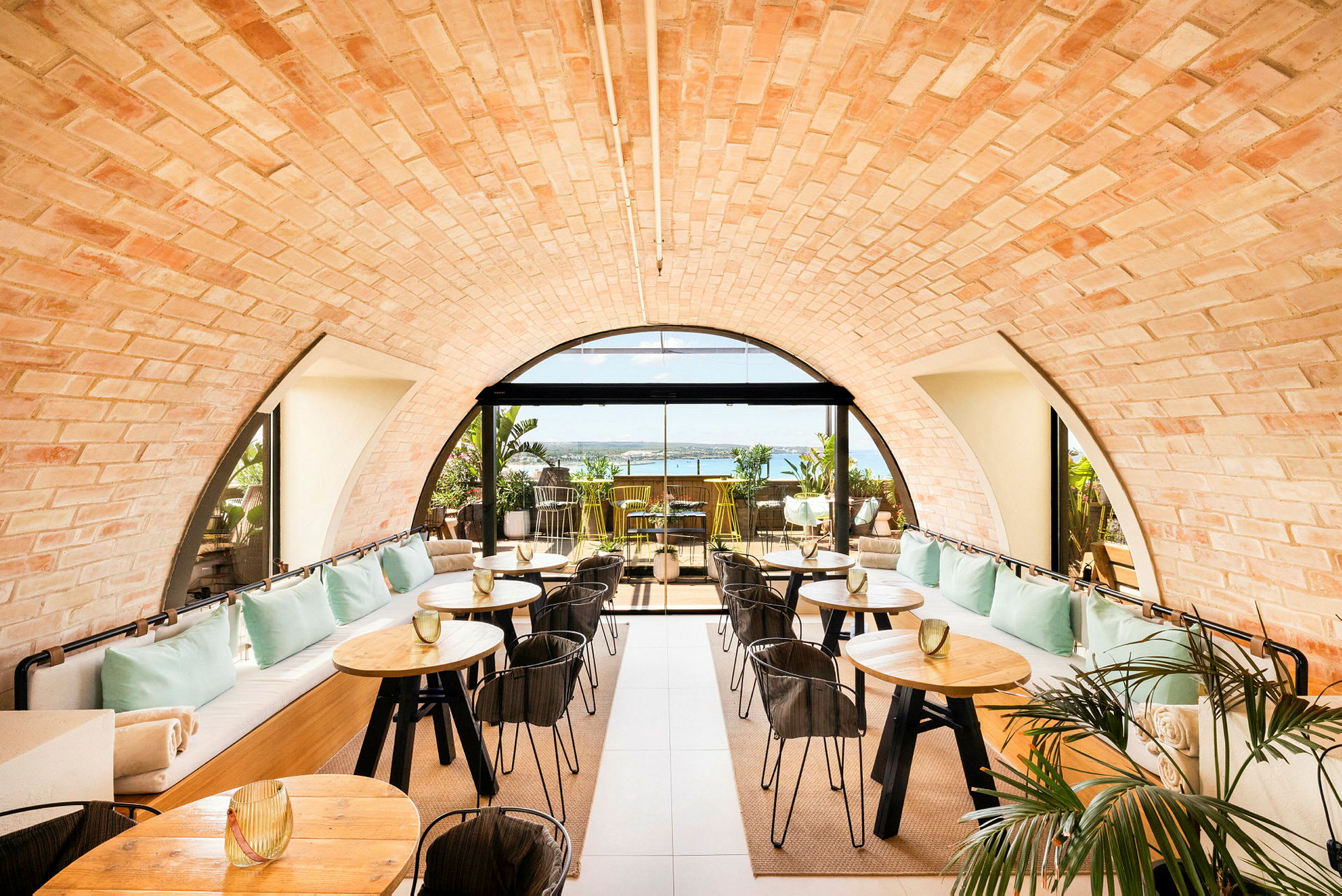 Et stilfuldt caféområde med buet loft af mursten og store vinduer, der åbner ud mod en terrasse med havudsigt. Bordene er af træ med sorte ben, og der er dekorative planter. (AI-genereret tekst)