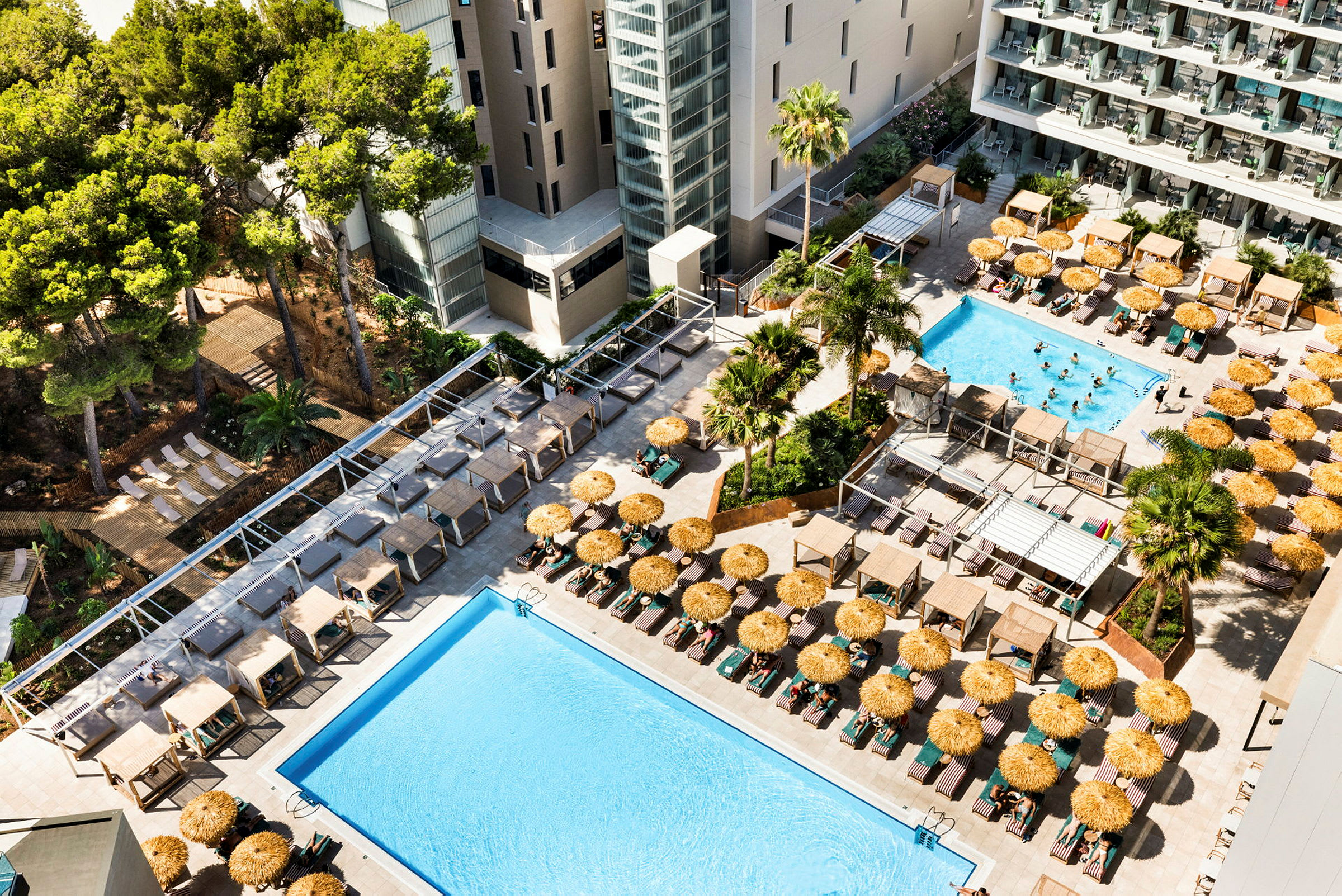 En hotelpool med liggestole og parasoller omgivet af bygninger og træer. (AI-genereret tekst)