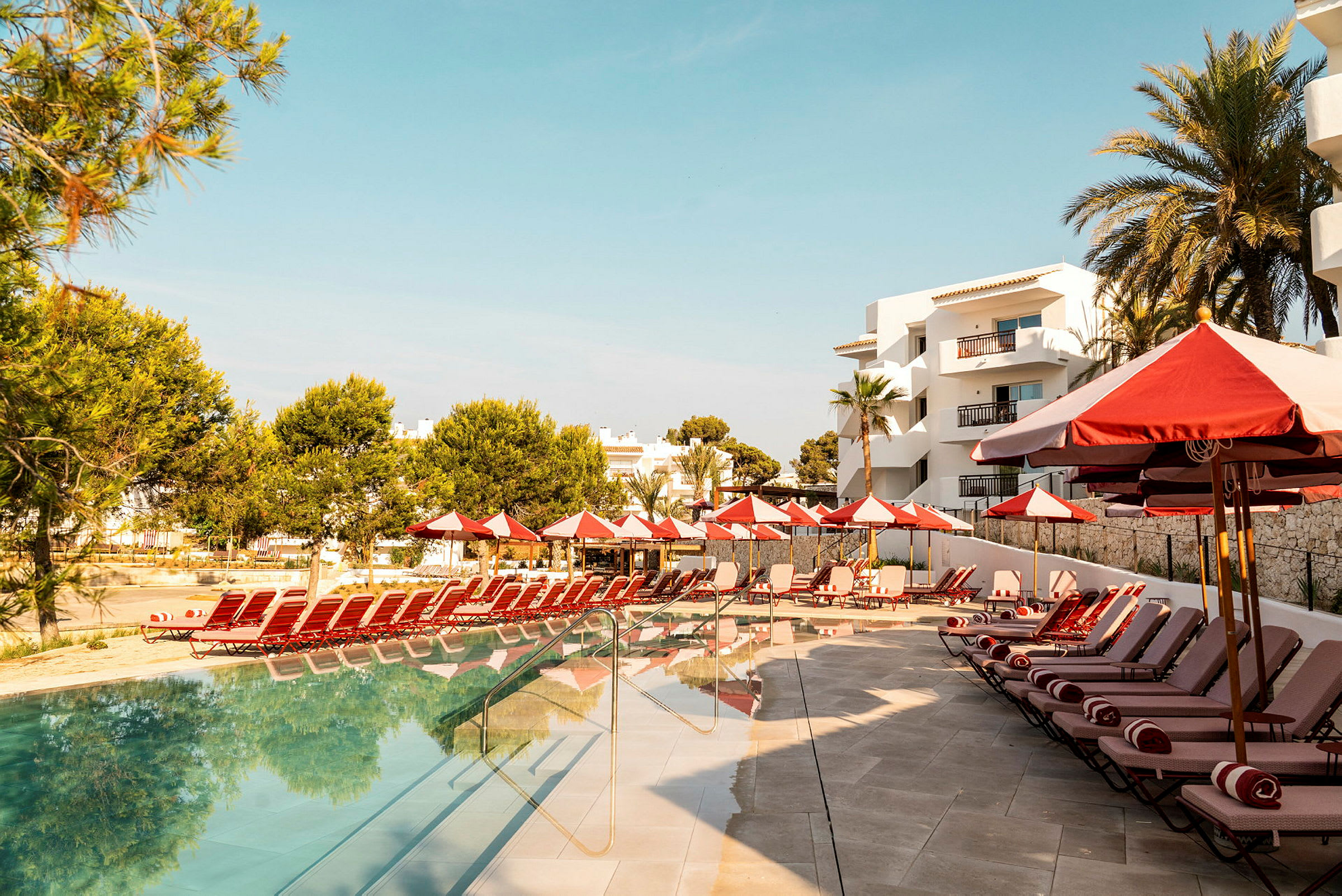 Billede af en hotelpool med liggestole og parasoller ved siden af bygninger. (AI-genereret tekst)
