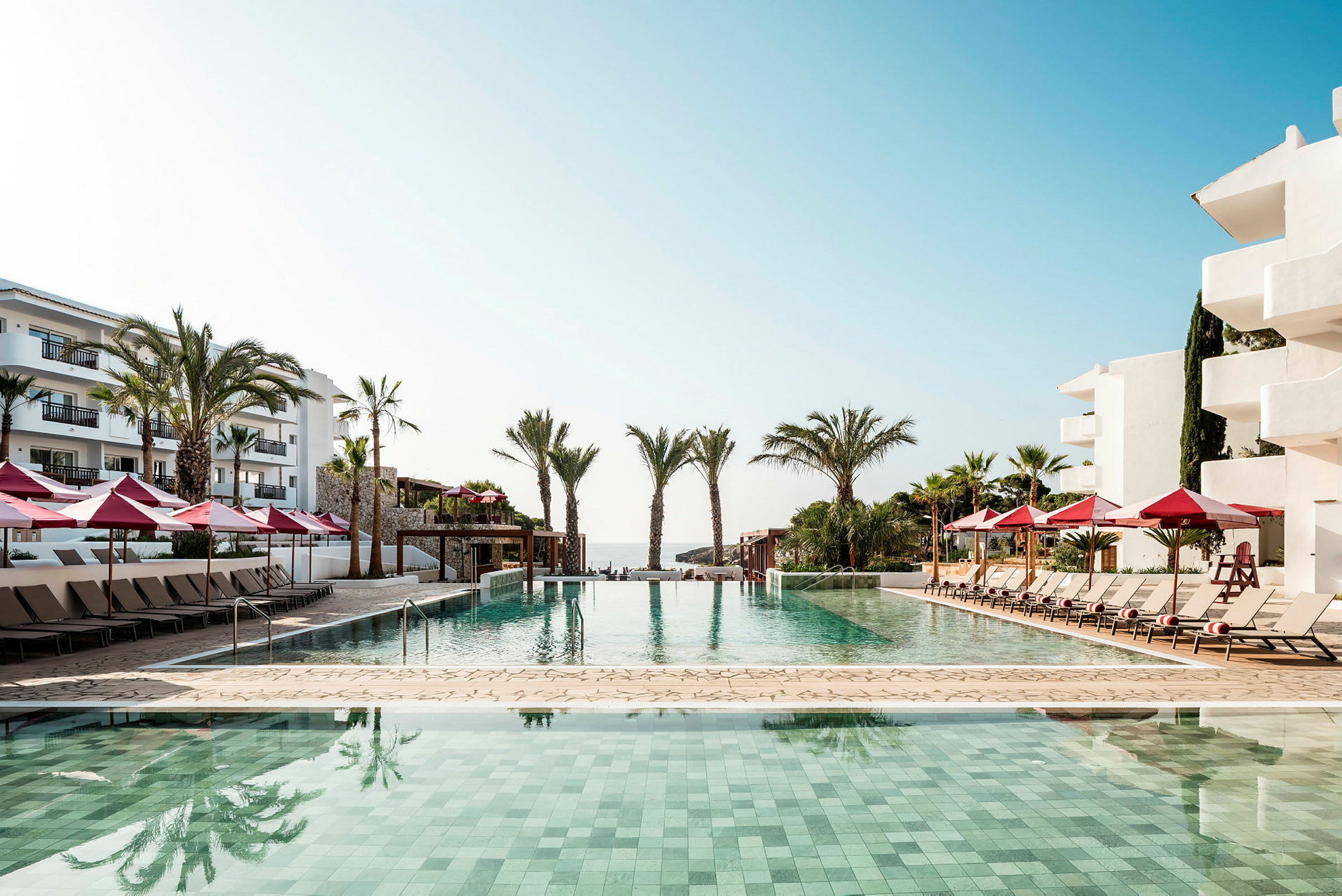 Udendørs pool ved kystnært hotel med liggestole og røde parasoller. (AI-genereret tekst)