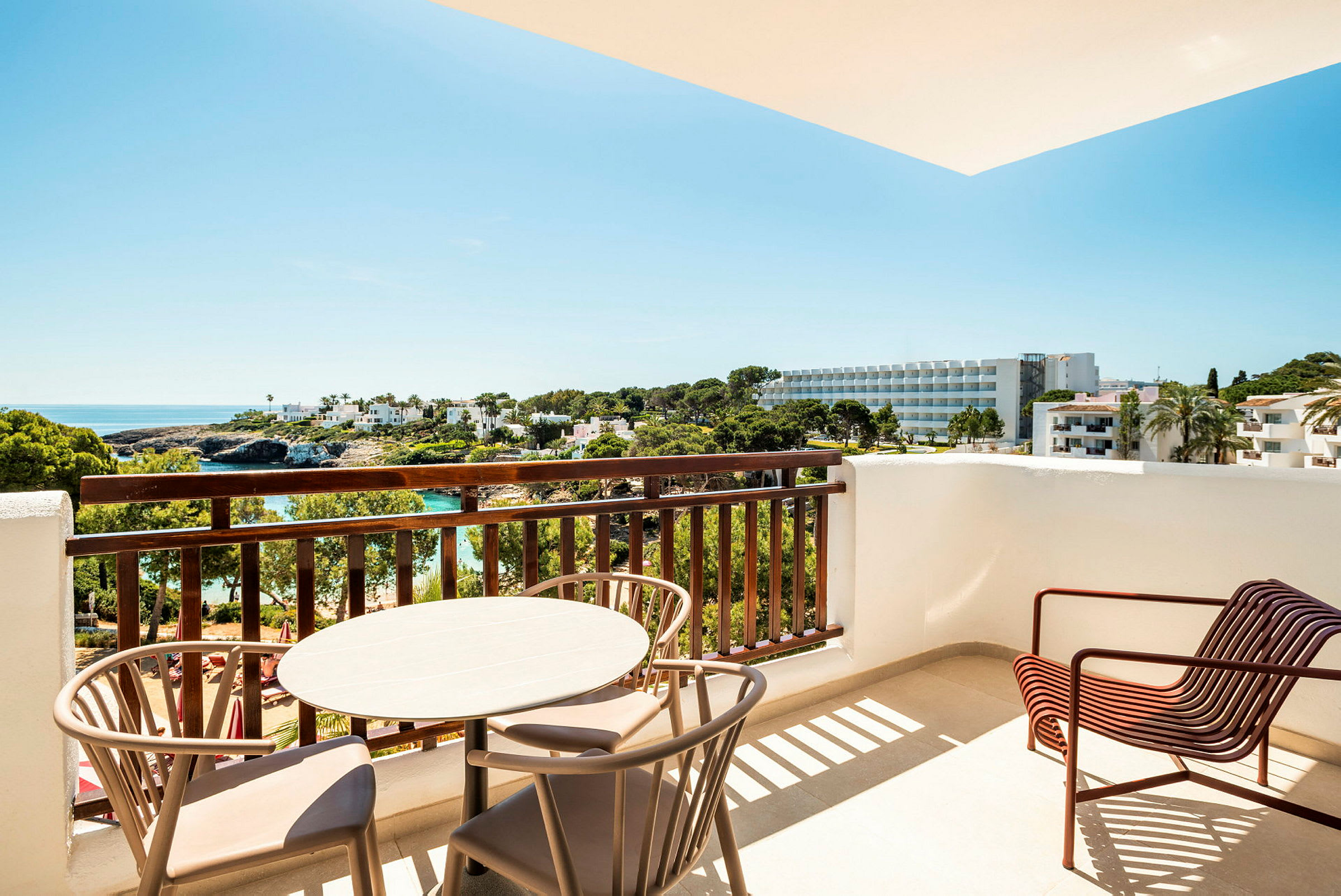 En balkon med et bord og stole, med udsigt over et område med træer, bygninger og havet i baggrunden. (AI-genereret tekst)