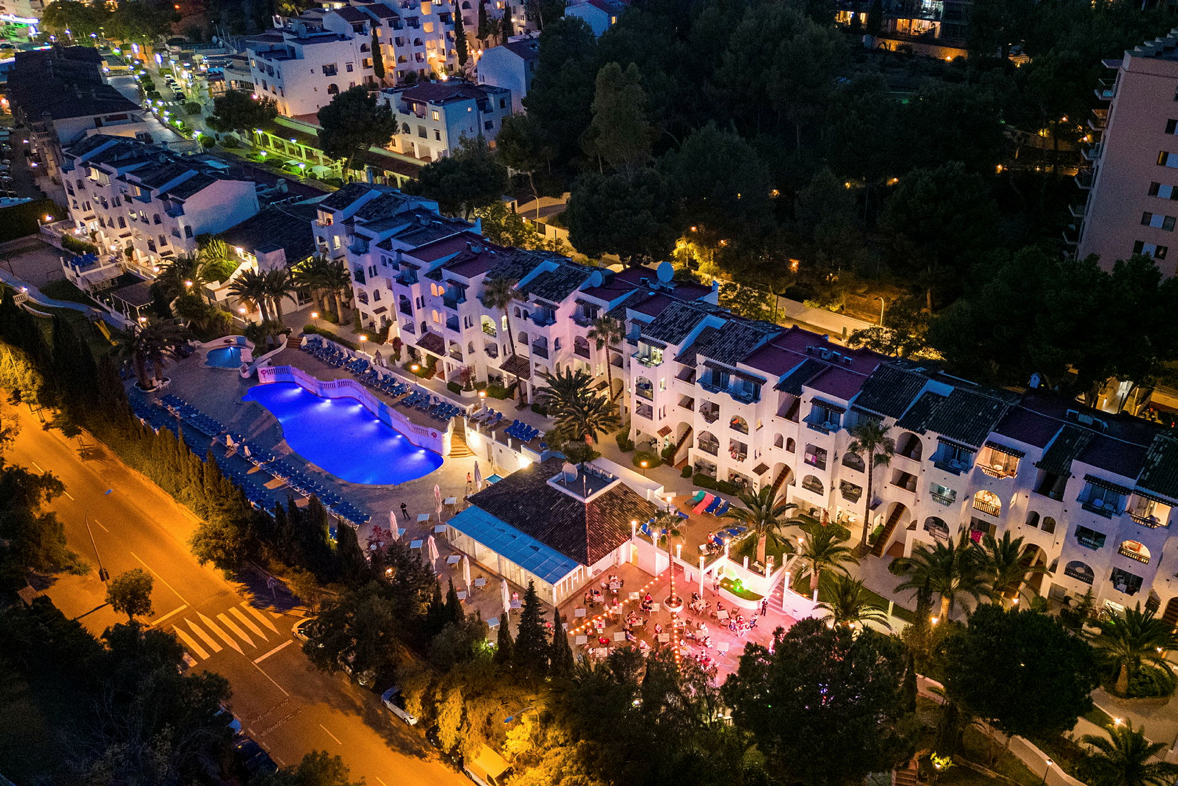 Luftfoto af en oplyst hotelanlæg med swimmingpool og terrasse om aftenen. (AI-genereret tekst)