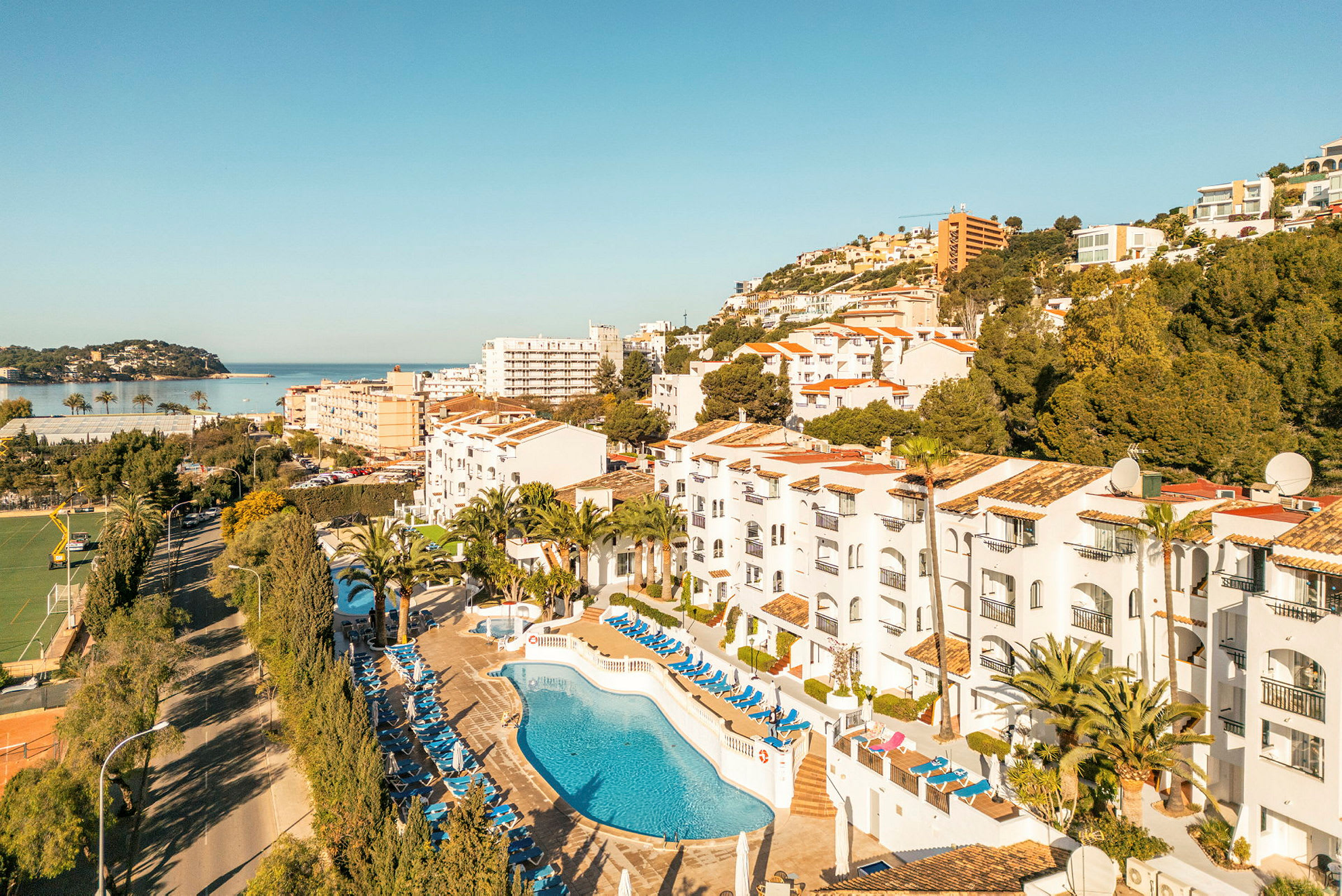 Billede af hotelområde med pool og liggestole, omgivelser med træer og bygninger samt udsigt mod havet. (AI-genereret tekst)
