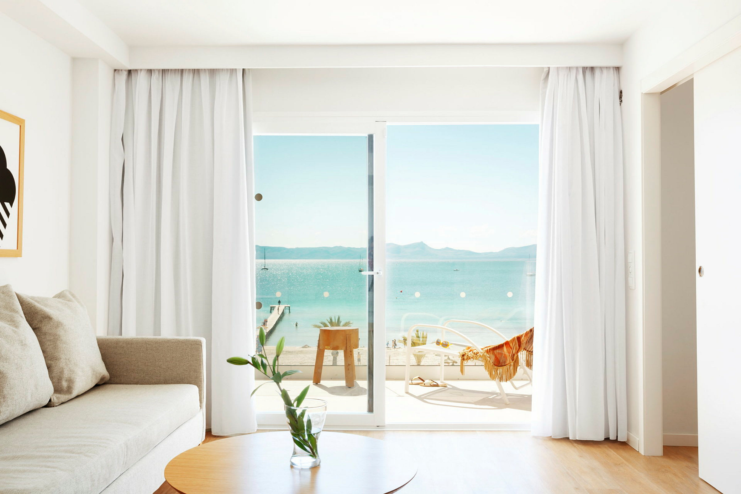 Billede af et lyst værelse med en sofa, et bord og glasdøre, der fører til en balkon med udsigt over havet. (AI-genereret tekst)