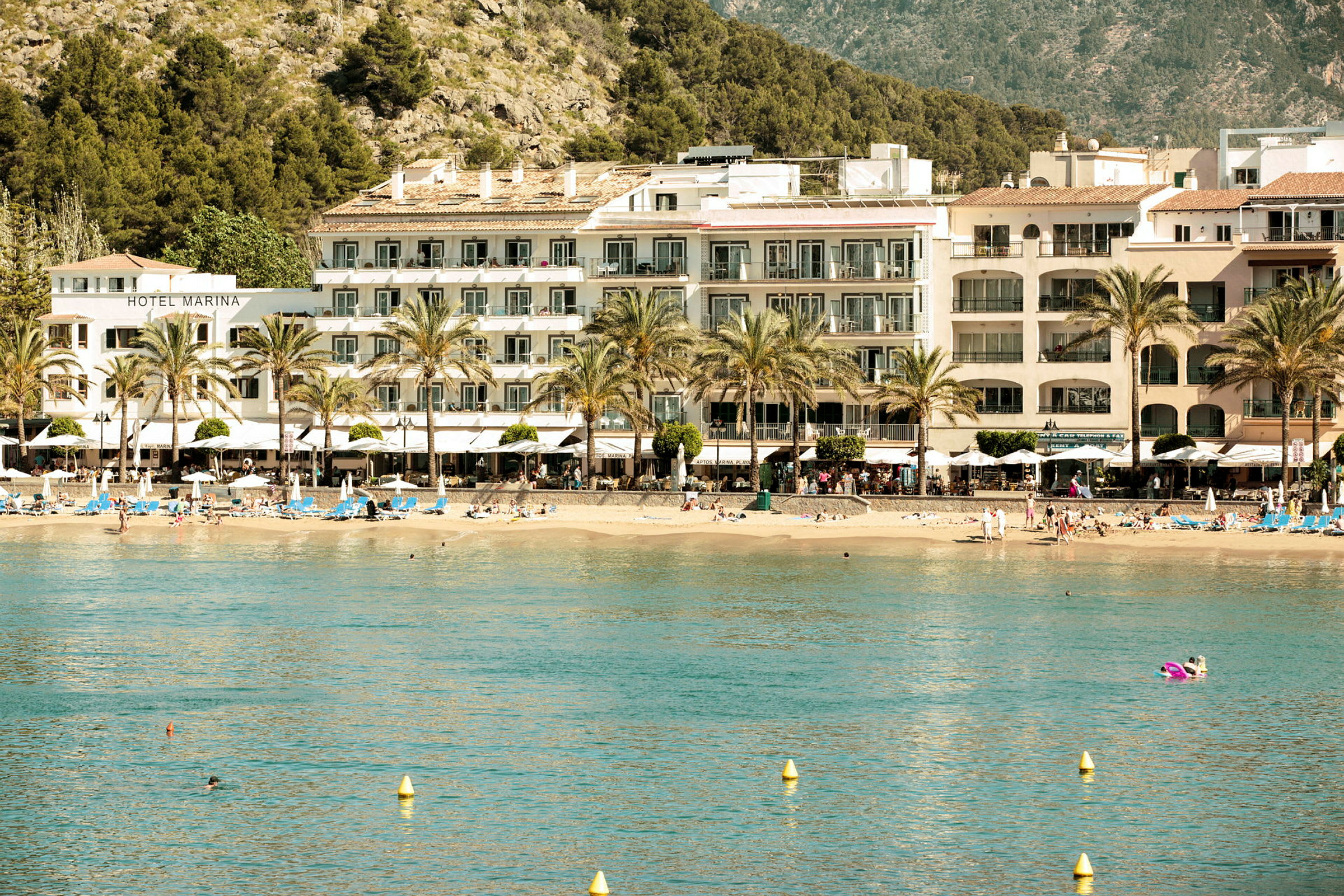 Hotel og strand ved kysten med bjerge og træer i baggrunden. (AI-genereret tekst)