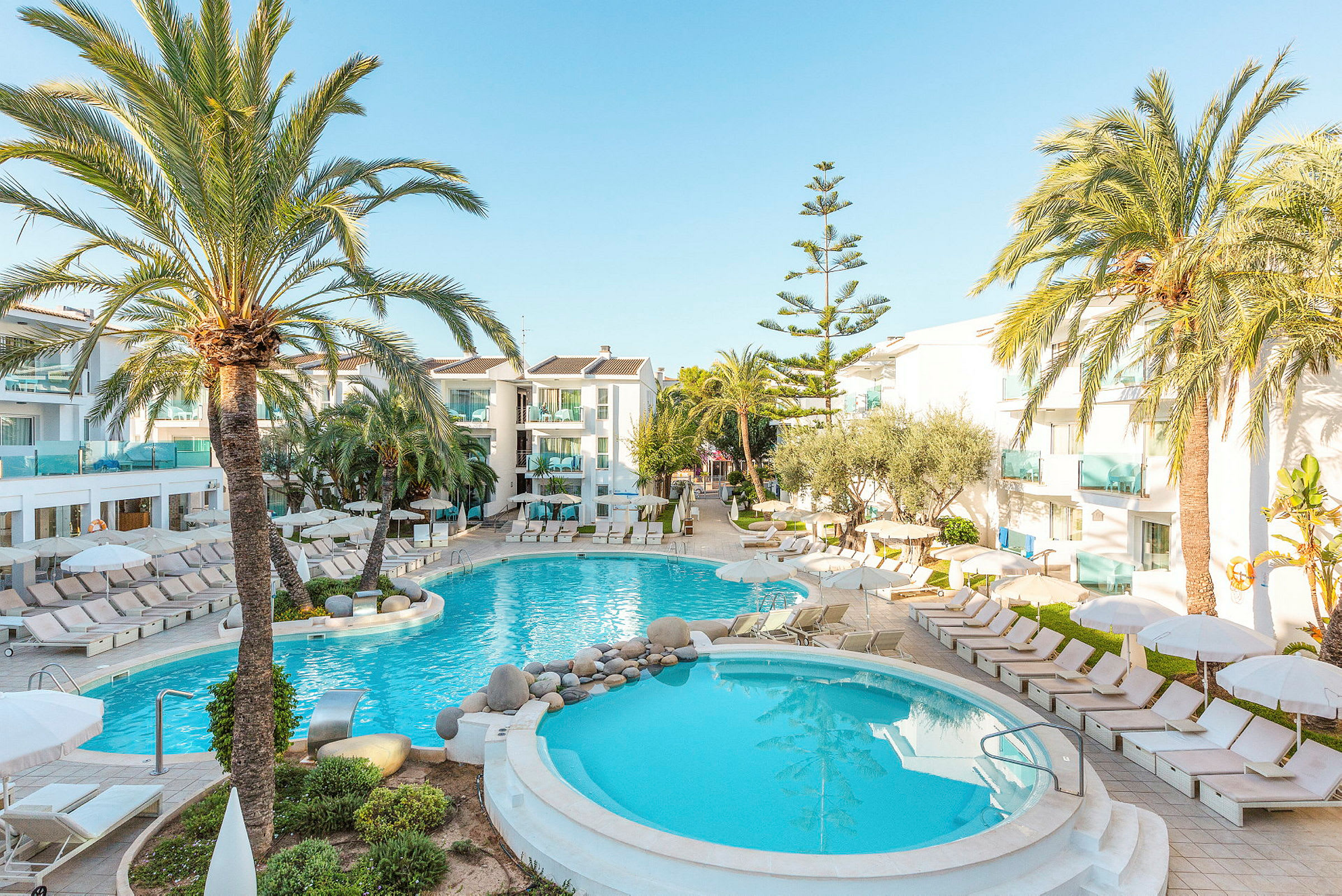 Billede af hotelområde med to swimmingpools omgivet af liggestole og palmer. (AI-genereret tekst)