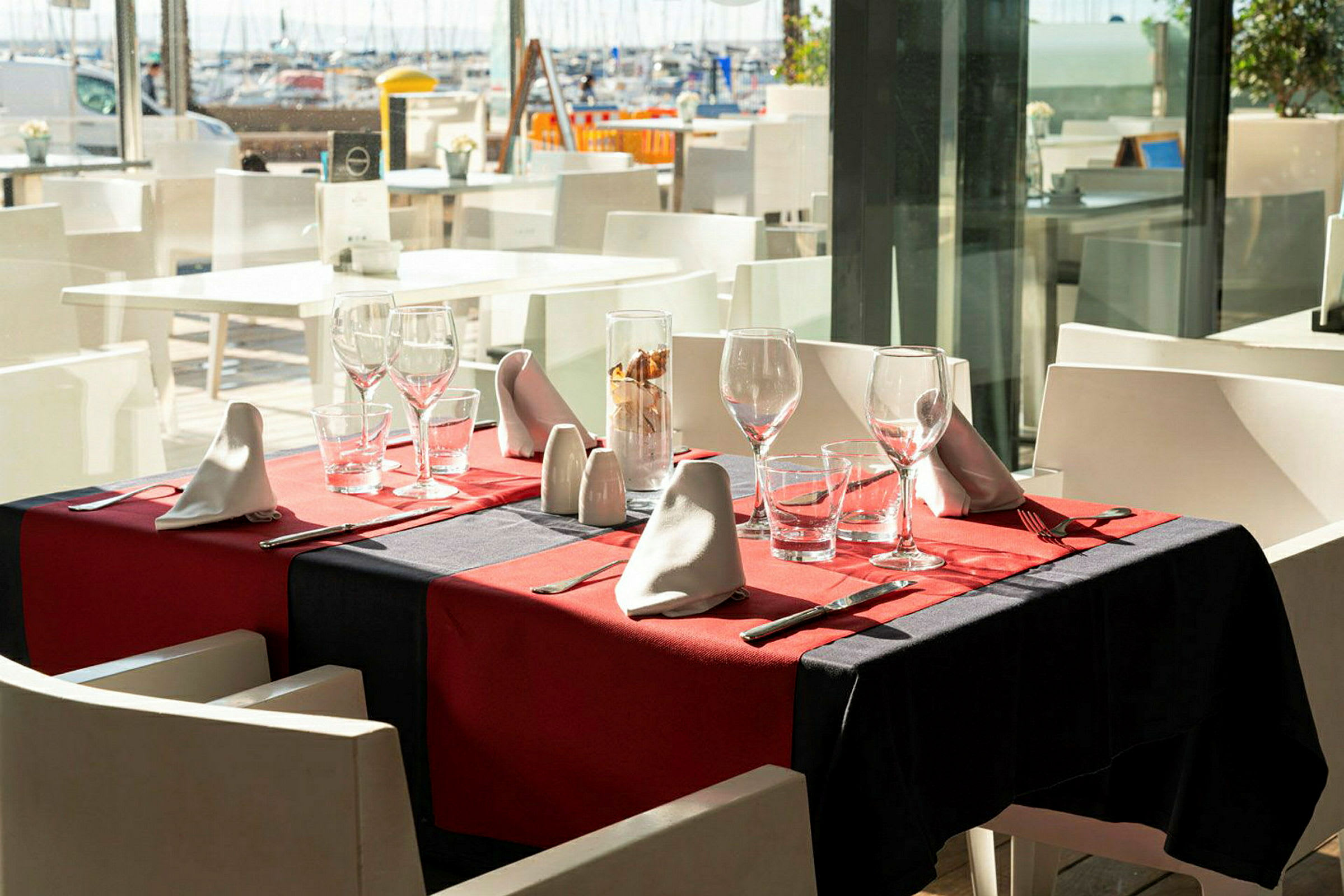Et dækket bord med rød dug, glas og bestik på en restaurant med udsigt over havnen. (AI-genereret tekst)