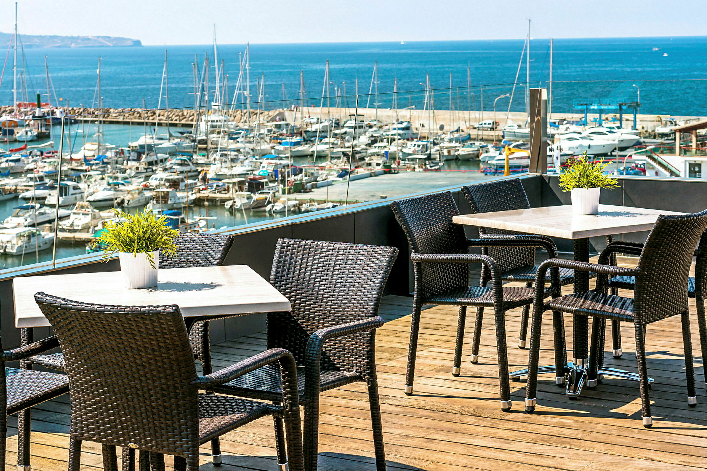Udendørs servering med borde og stole på en træterrasse, med udsigt over en lystbådehavn og havet. (AI-genereret tekst)