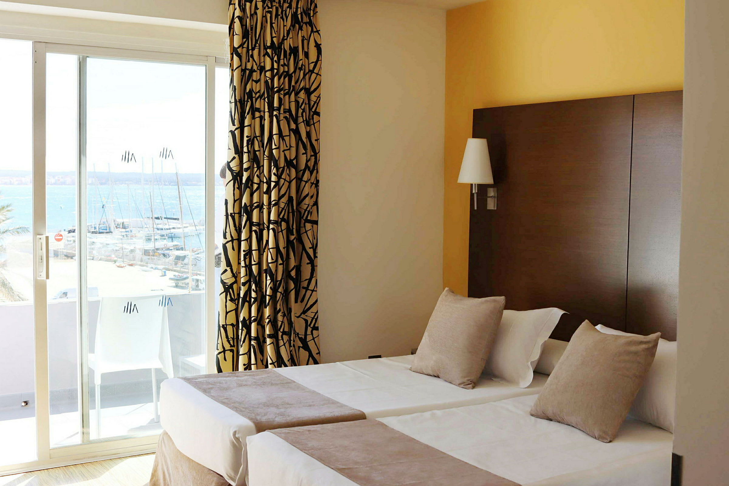 Hotelværelse med to enkeltsenge, dekoreret med puder og sengetæpper. Et vindue med skydedør viser udsigt over en marina. (AI-genereret tekst)