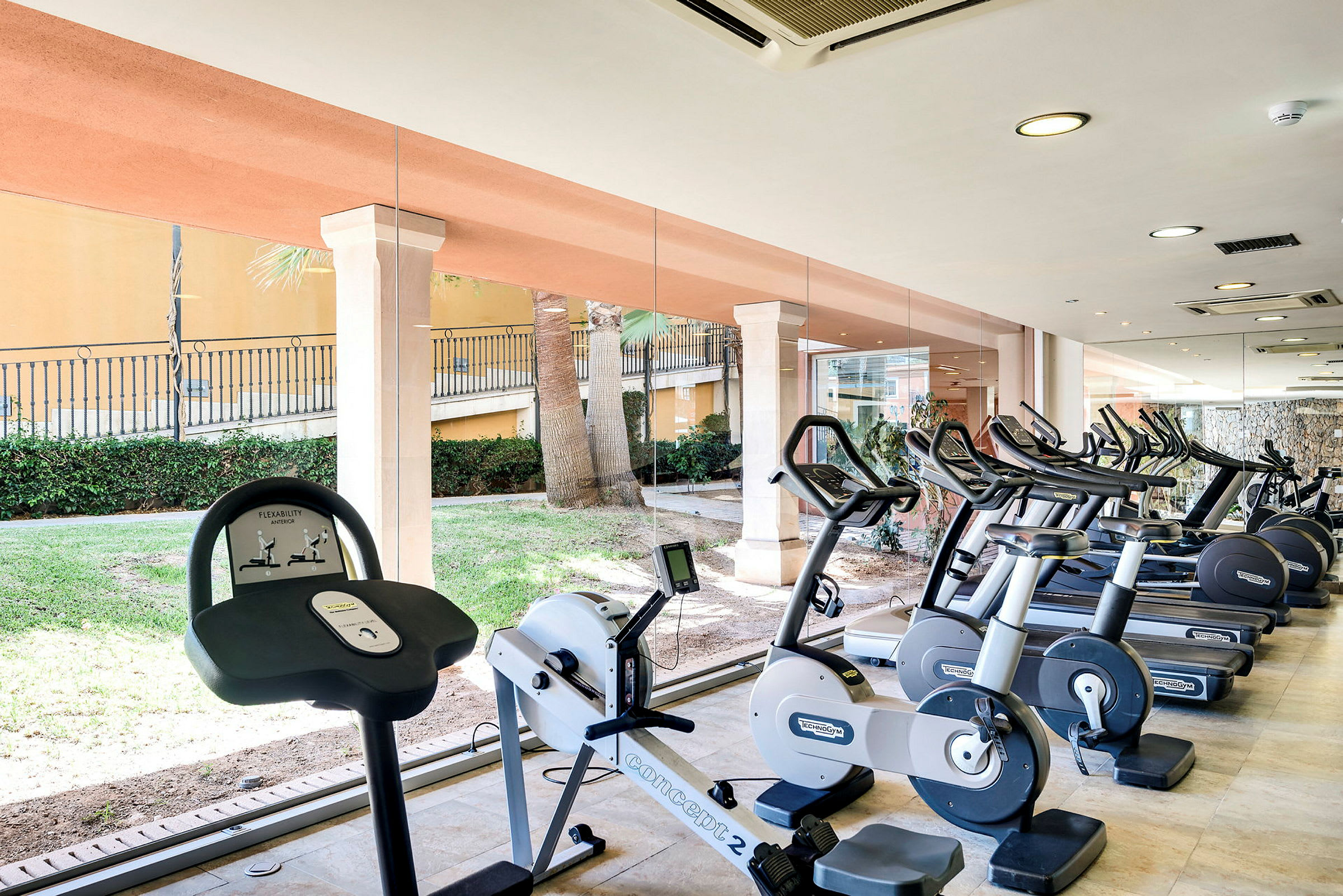 Billede af et fitnesscenter med træningsmaskiner, herunder cykler og løbebånd, med glasvægge der vender ud mod et grønt område. (AI-genereret tekst)