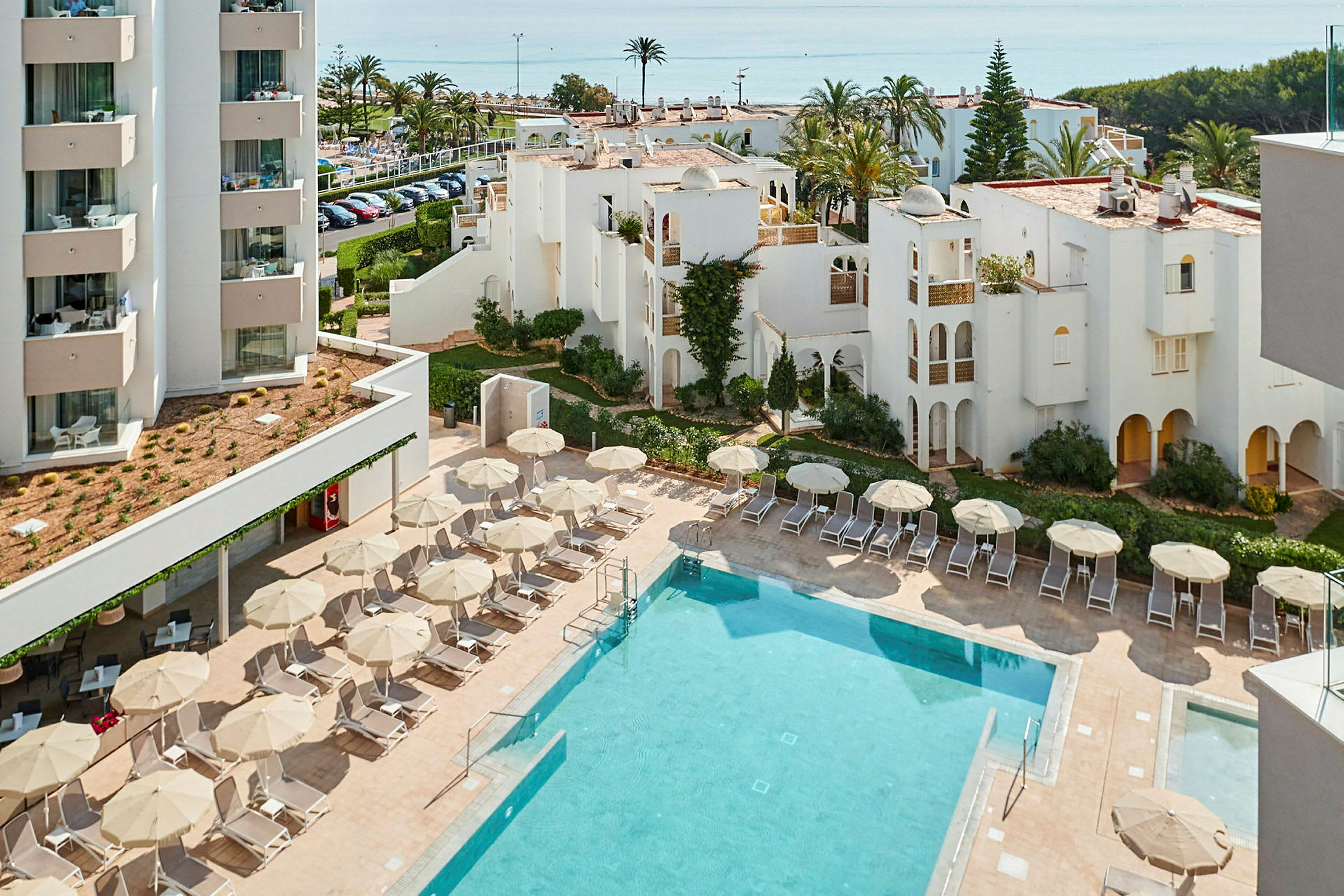 Hotelområdet med pool, liggestole og parasoller samt hvide bygninger i baggrunden. (AI-genereret tekst)