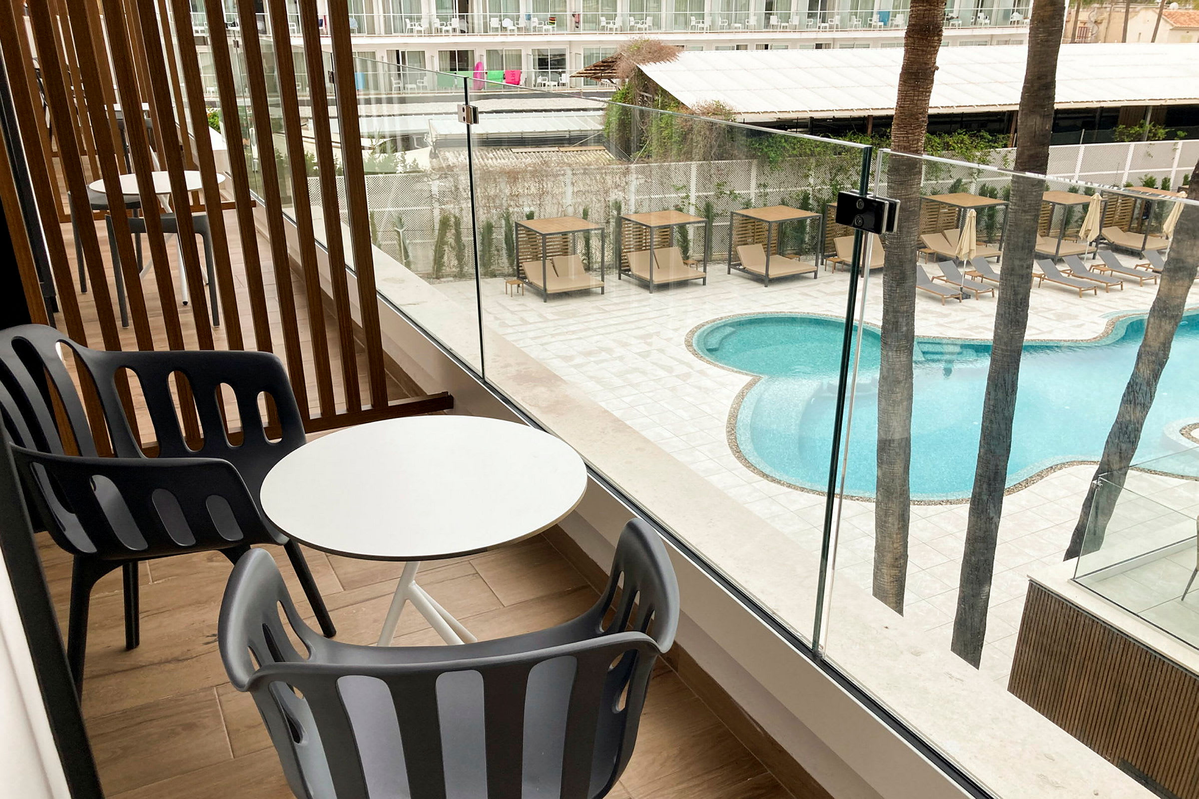 Billede af en balkon med bord og stole, med udsigt til en pool og solsenge. (AI-genereret tekst)