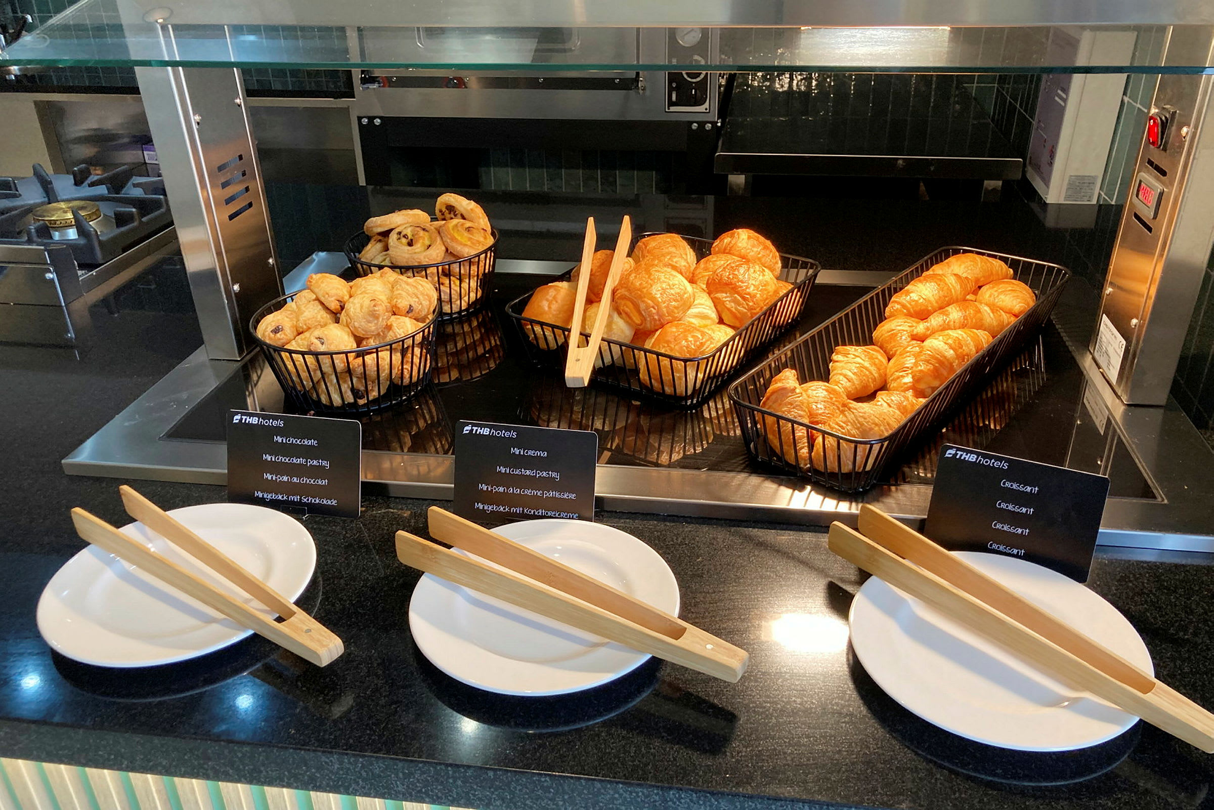 Morgenbuffet med friske croissanter og bagværk i sorte kurve. Foran ligger hvide tallerkener med trætænger. (AI-genereret tekst)