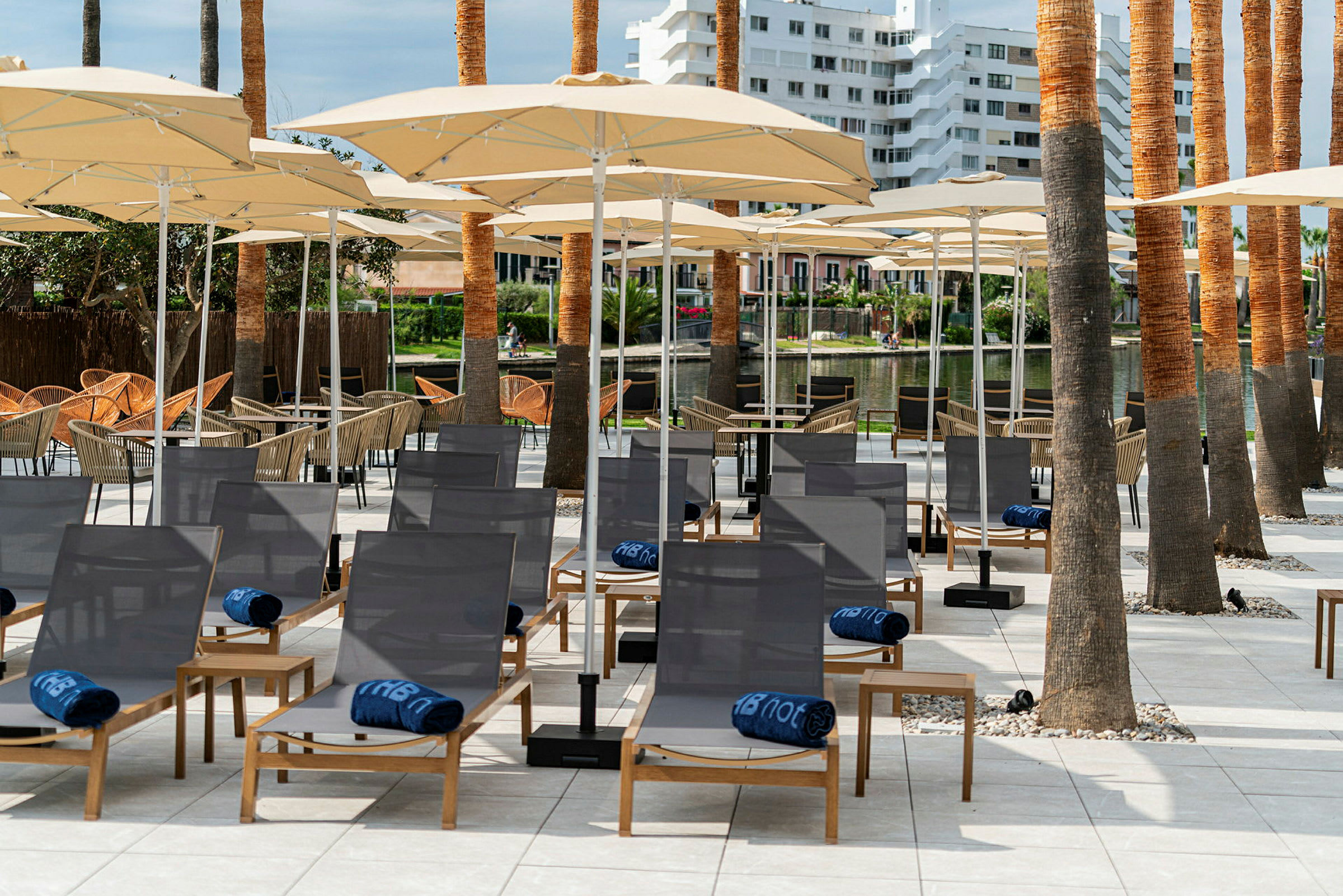 Liggestole med blå håndklæder og parasoller på en terrasse omgivet af palmer. (AI-genereret tekst)