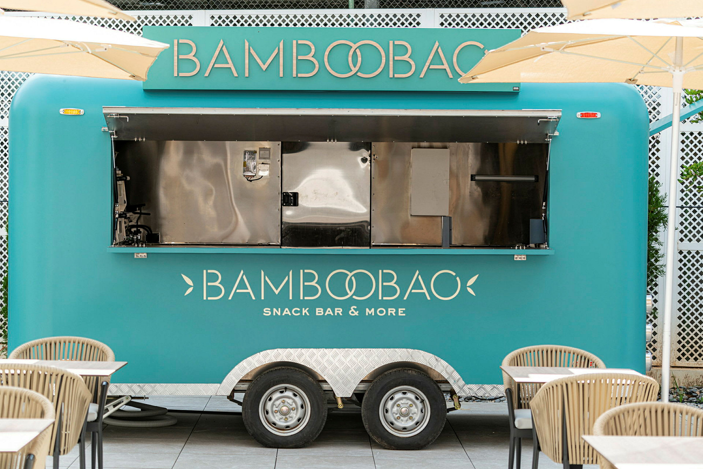 En turkis madvogn med teksten 'Bamboobao Snack Bar & More' står parkeret omgivet af borde og stole. (AI-genereret tekst)