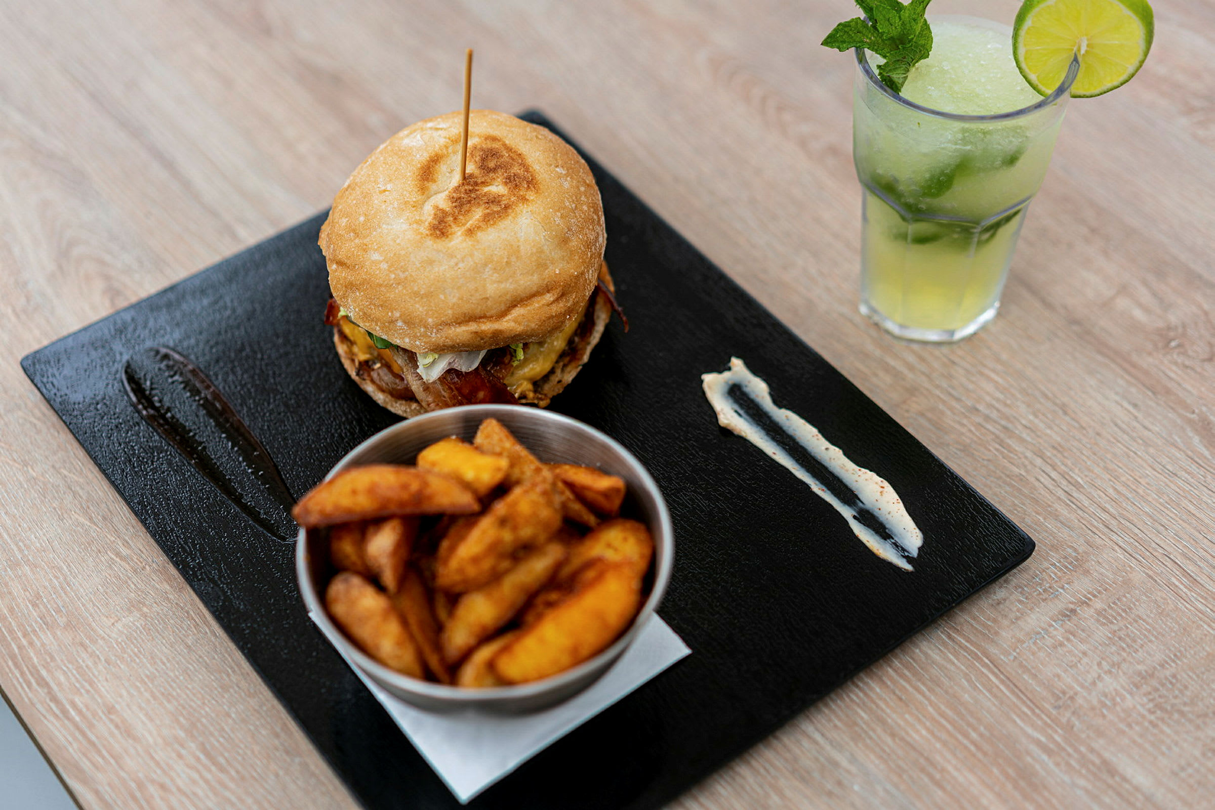 En tallerken med en burger, en skål med kartoffelbåde og en forfriskende drink med lime og mynte. (AI-genereret tekst)