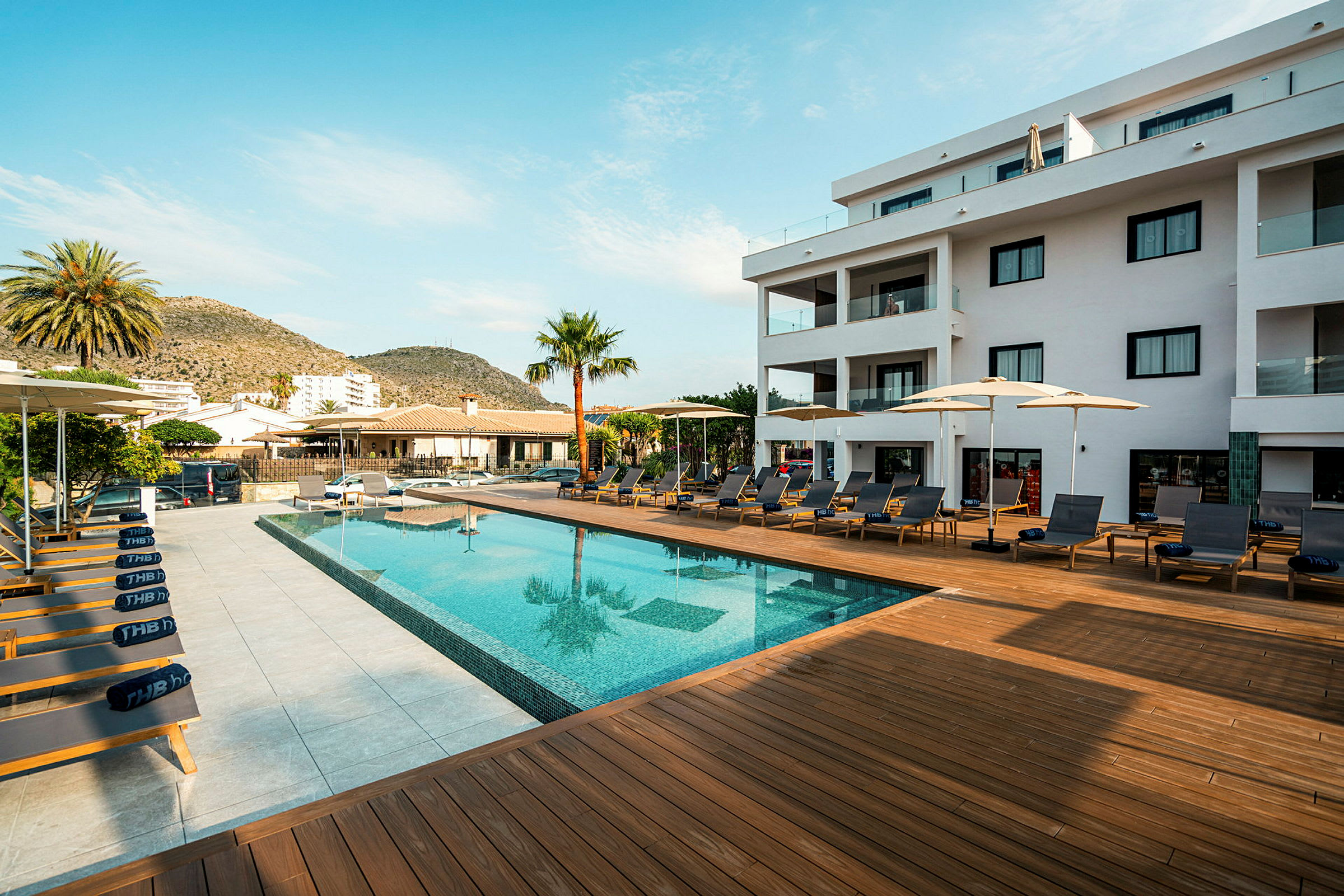 En udendørs swimmingpool med liggestole og parasoller omgivet af terrassebrædder og en moderne bygning i baggrunden. (AI-genereret tekst)