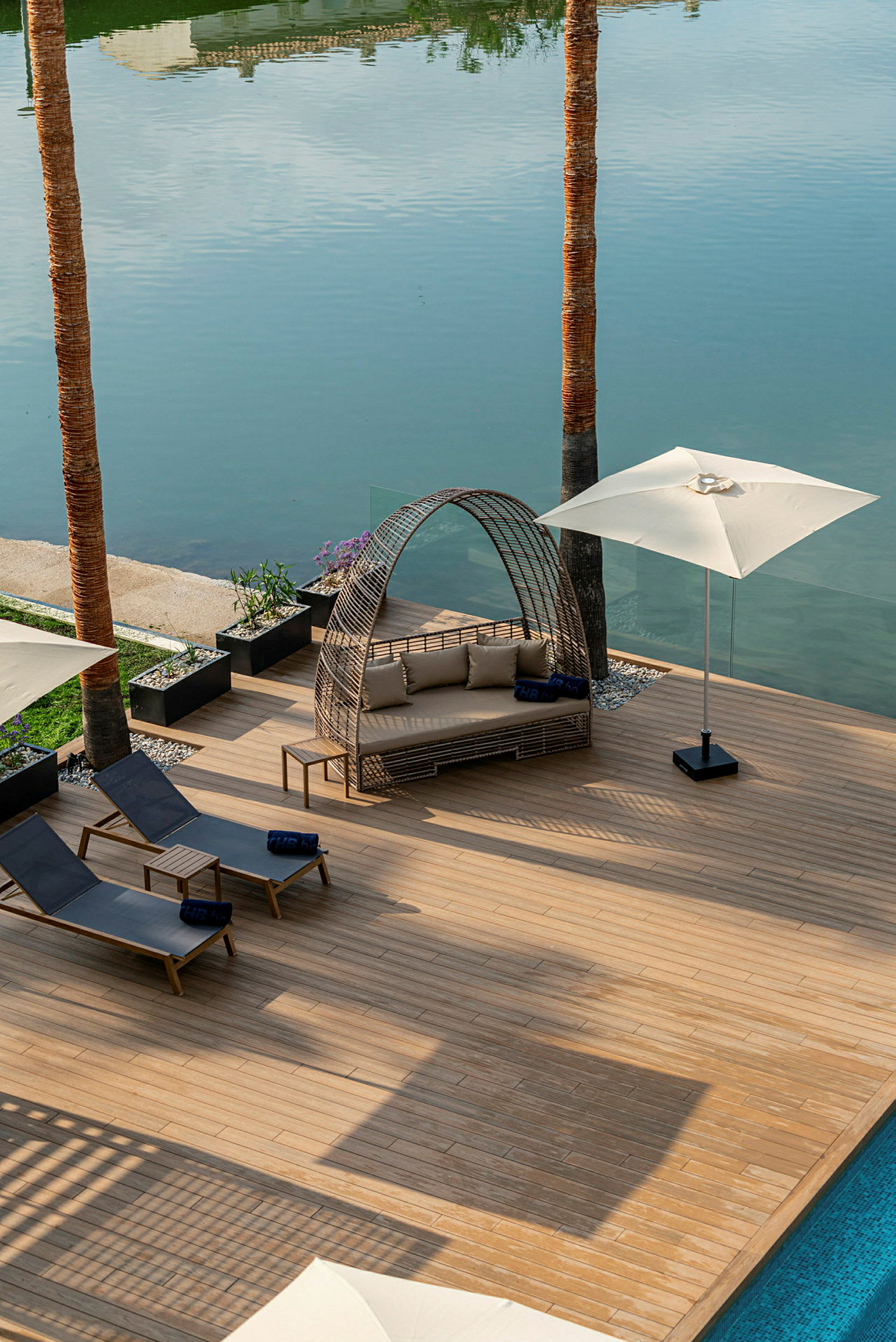 En terrasse med liggestole, parasol og en siddeplads ved siden af en pool og en sø. (AI-genereret tekst)