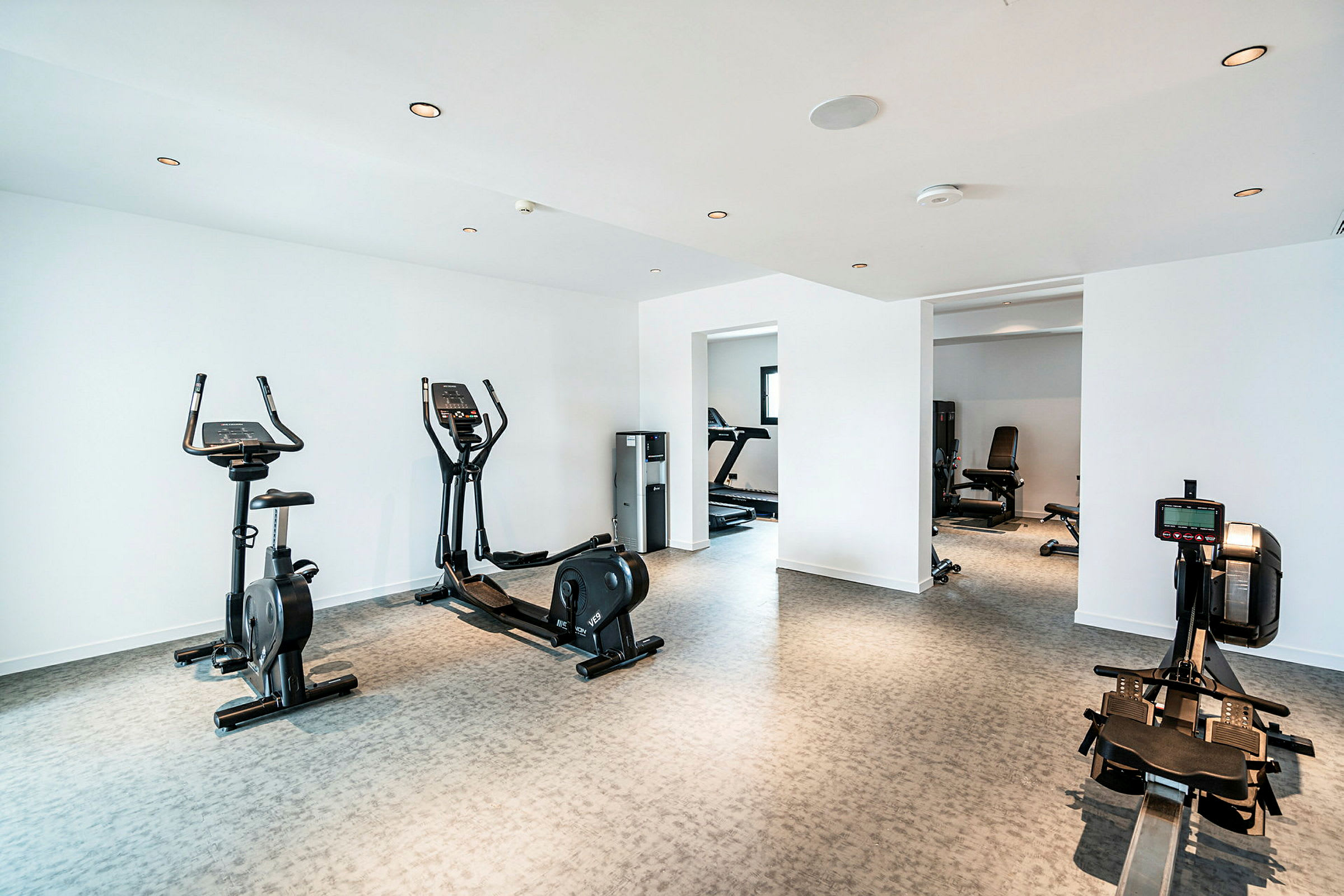 Et lyst fitnessrum med træningsudstyr såsom kondicykel, romaskine og crosstrainer samt vinduer og andet træningsudstyr i baggrunden. (AI-genereret tekst)