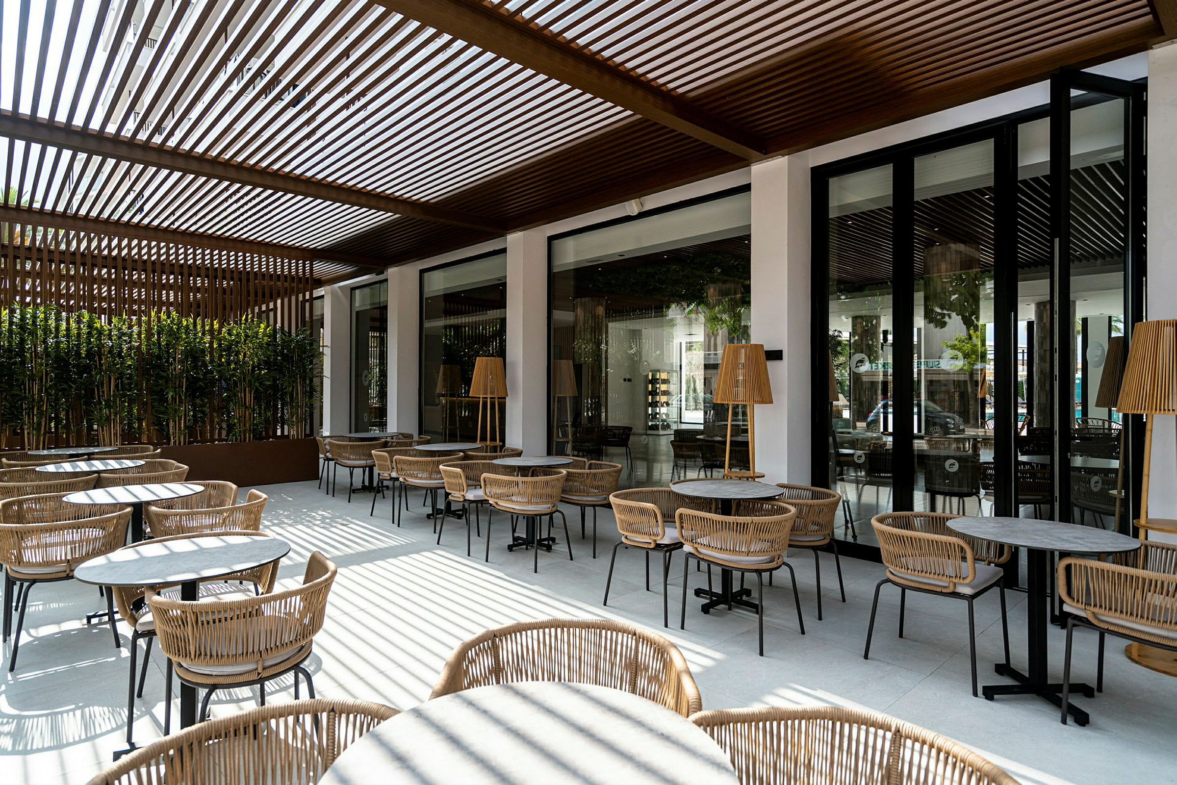 En udendørs serveringsområde med borde og stole, omgivet af store vinduer og en skyggefuld pergola. (AI-genereret tekst)