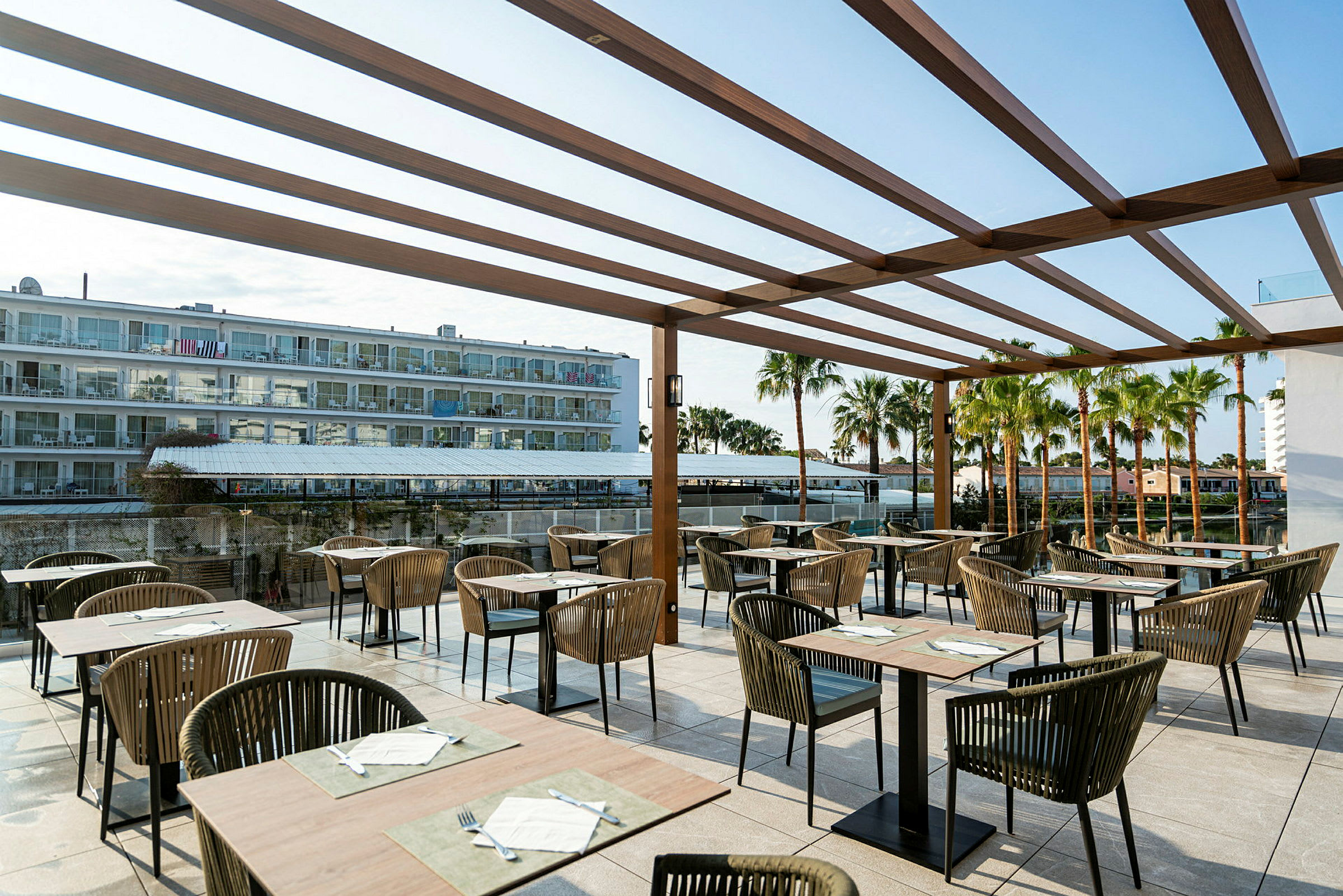 Billede af en restaurantterrasse med borde og stole under en træpergola. I baggrunden ses et hotel og palmer. (AI-genereret tekst)
