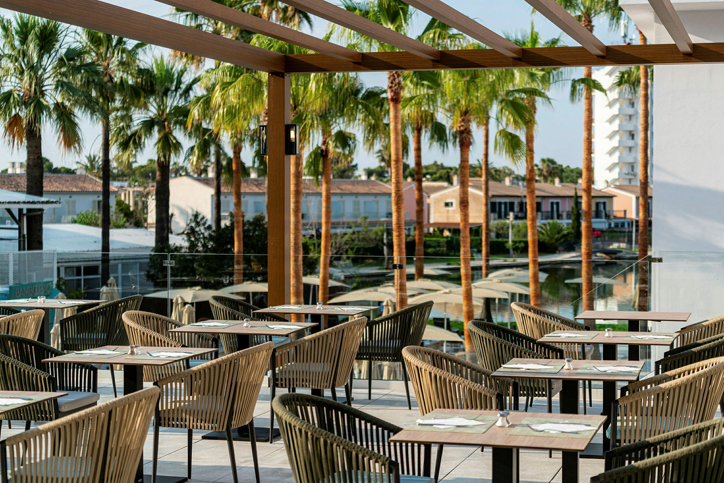 En udendørs terrasse med borde og stole under en pergola, omgivet af palmer og bygninger i baggrunden. (AI-genereret tekst)