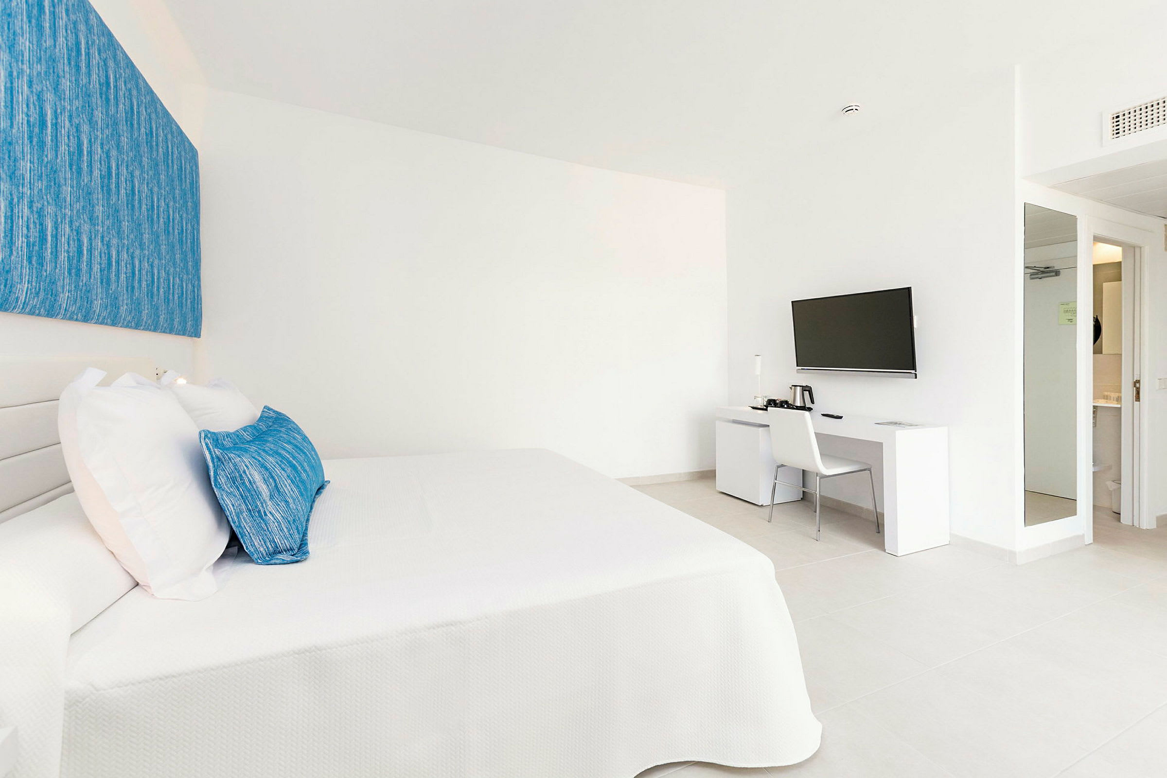 Et hotelværelse med en hvid seng med blå puder og et tilstødende skrivebord med et væghængt tv over. (AI-genereret tekst)