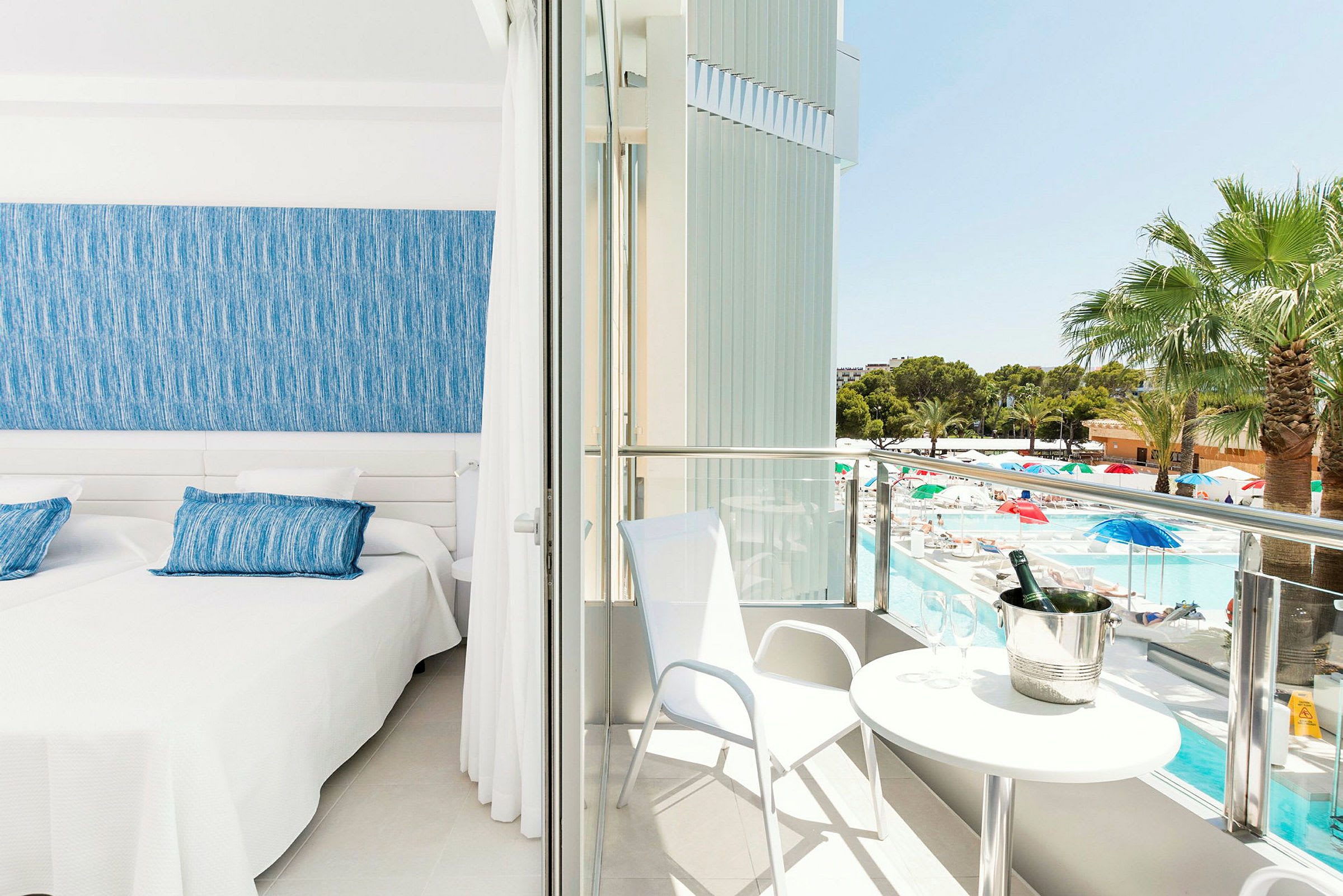 Et hotelværelse med to senge, et blåt sengegærde og adgang til en balkon med udsigt over en pool og have. (AI-genereret tekst)