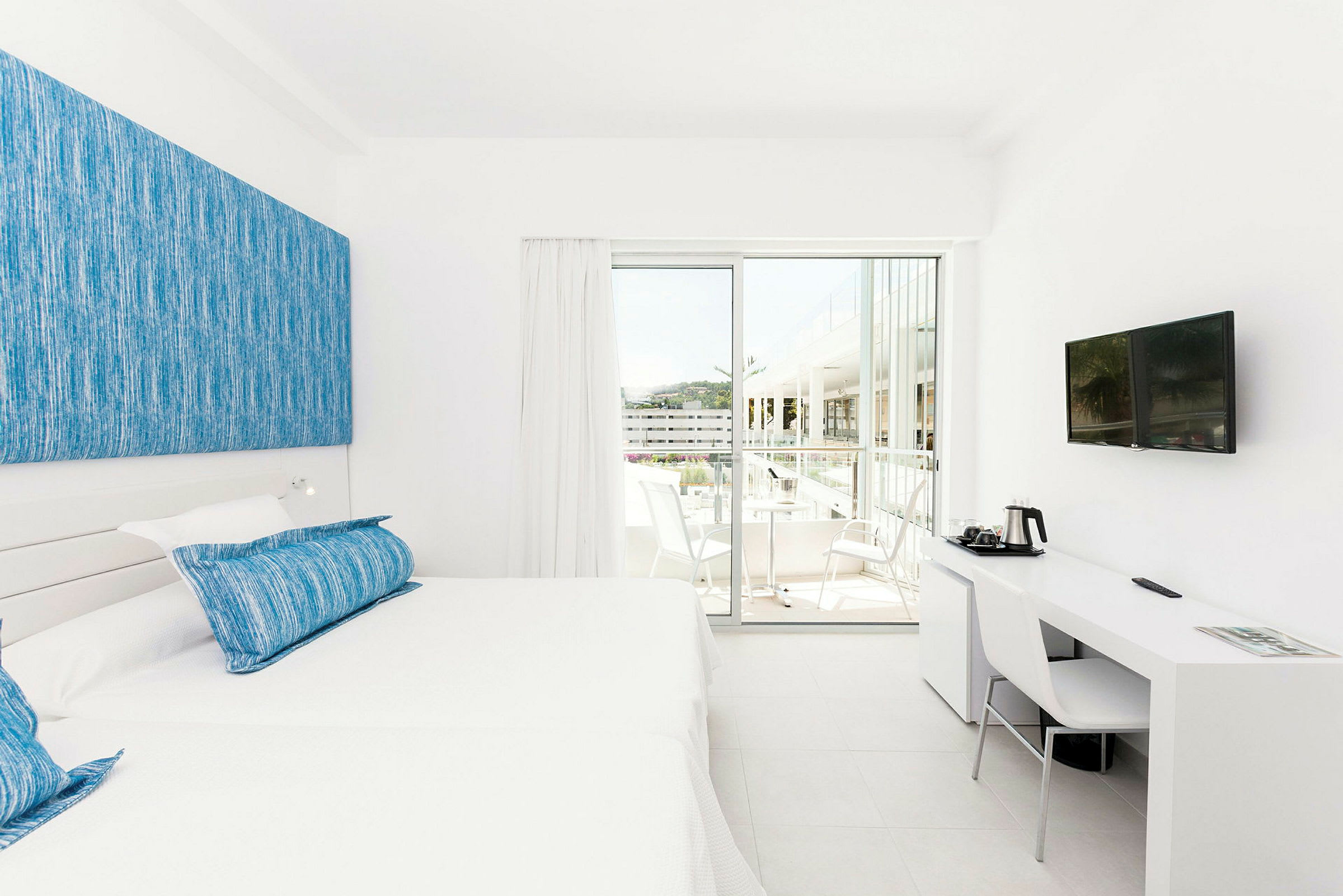 Et lyst hotelværelse med hvidt interiør, to enkeltsenge med blå puder og et skrivebord med en stol og et TV på væggen. En skydedør fører ud til en balkon med siddepladser. (AI-genereret tekst)
