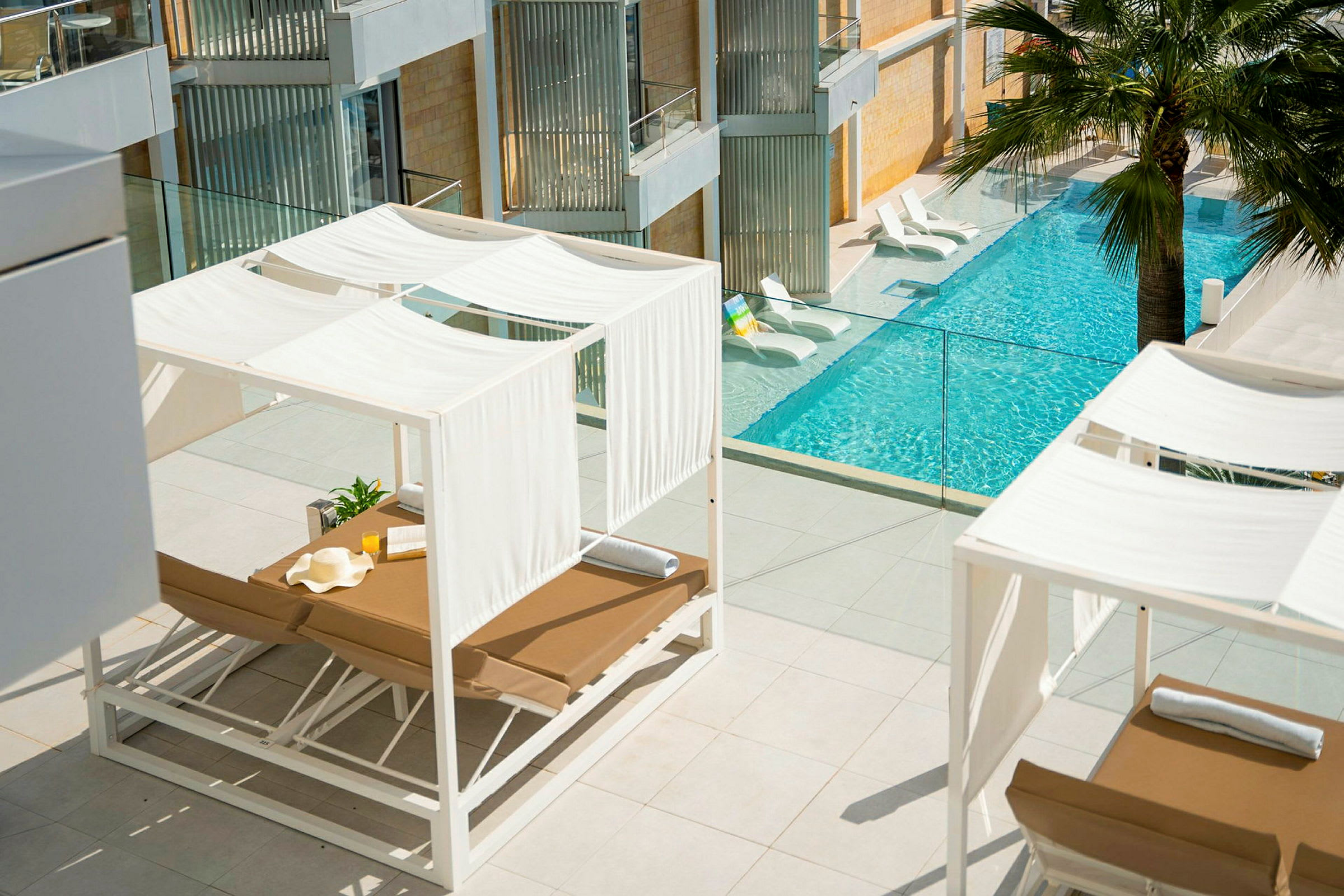 En terrasse med hvide liggestole under skyggefulde baldakiner og udsigt over en pool med klart vand. (AI-genereret tekst)
