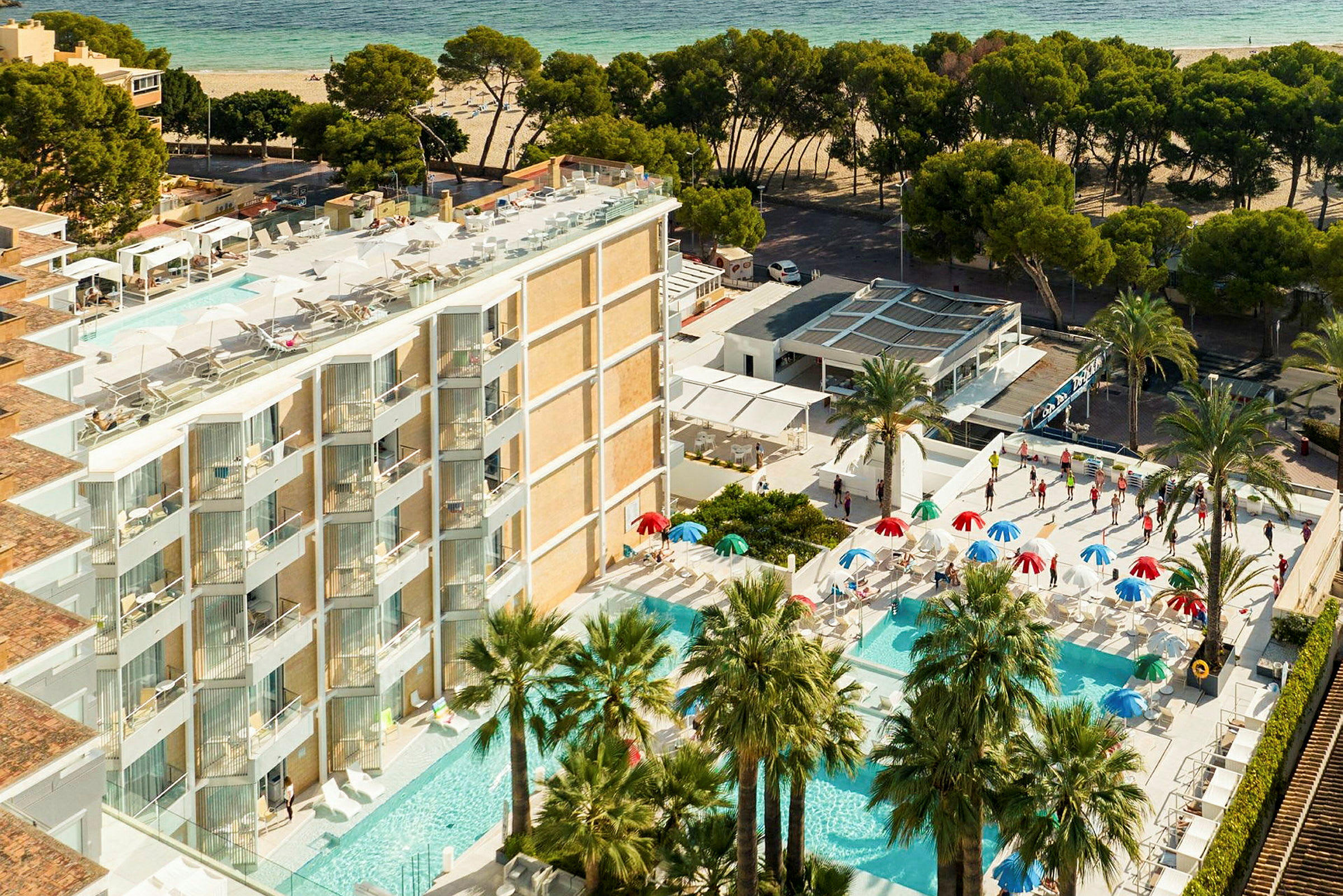 Udsigt over hotelområde med flere pools, parasoller og palmer tæt på en sandstrand. (AI-genereret tekst)
