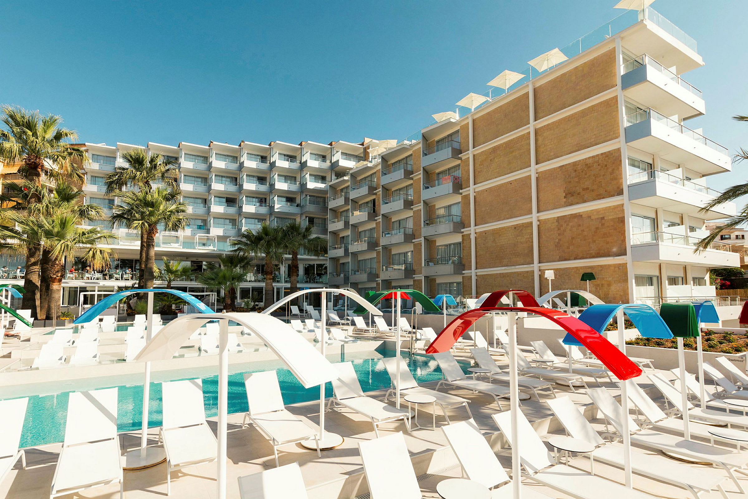 Et hotel med flere etager i moderne design kan ses bag et poolområde med liggestole og farverige solskærme. (AI-genereret tekst)