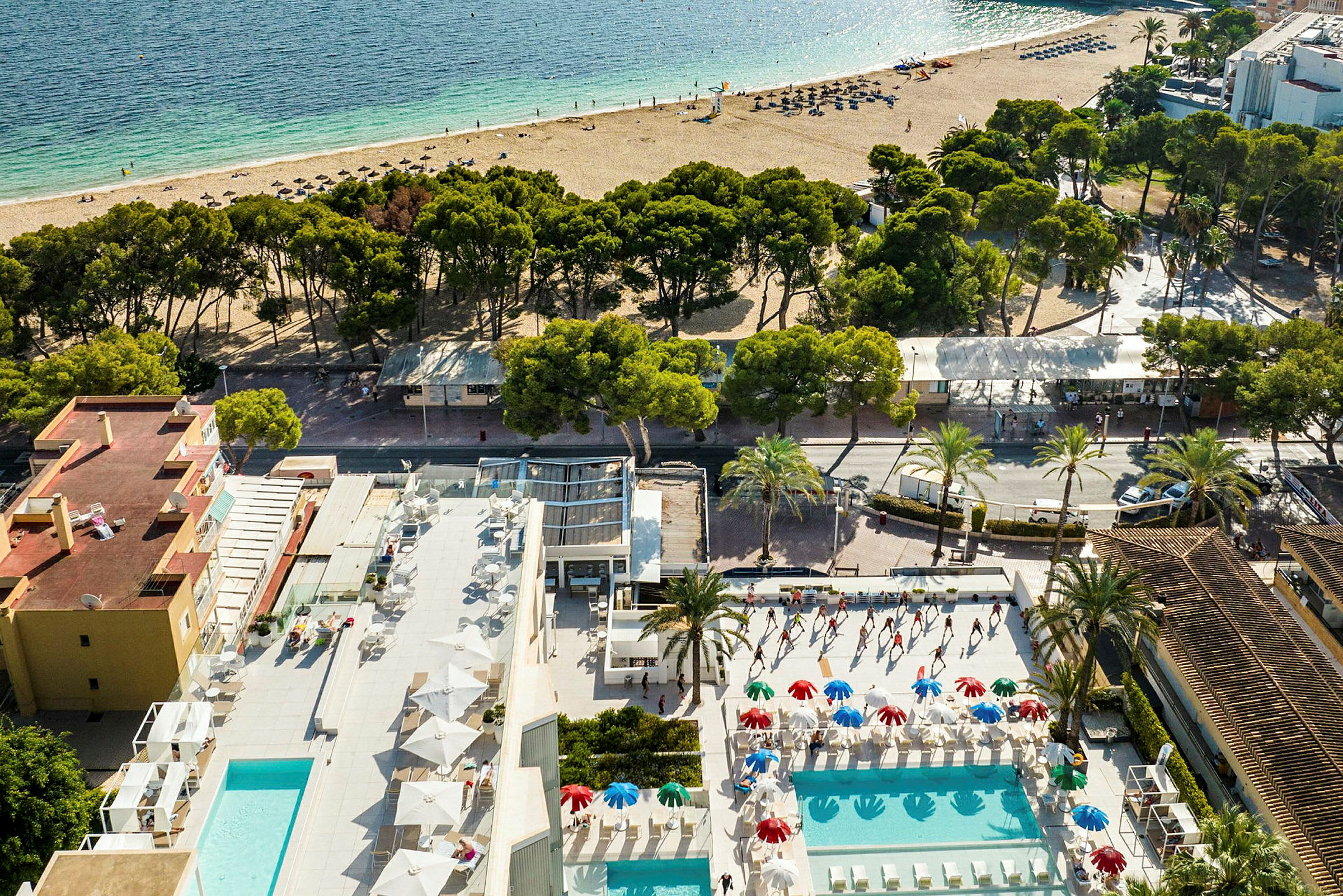 Udsigt fra oven af et hotelområde med udendørs swimmingpools, parasoller og liggestole. En sandstrand og havet ses i baggrunden. (AI-genereret tekst)