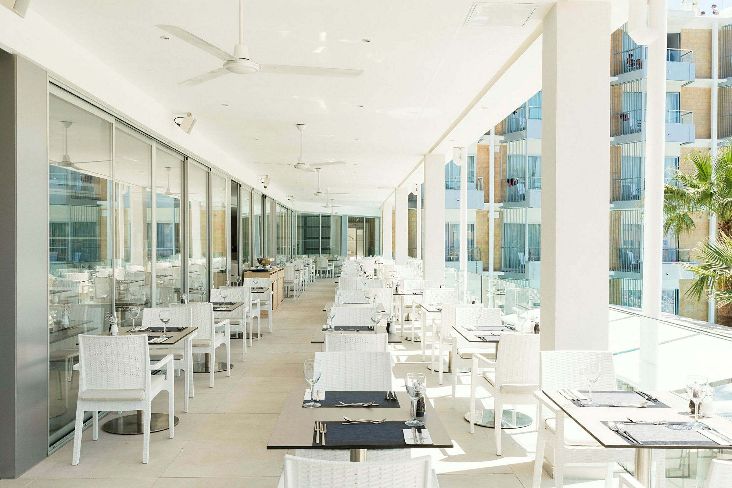 En lys restaurantterrasse med mange hvide borde og stole, glasdøre langs den ene side og udsigt over altaner udenfor. (AI-genereret tekst)