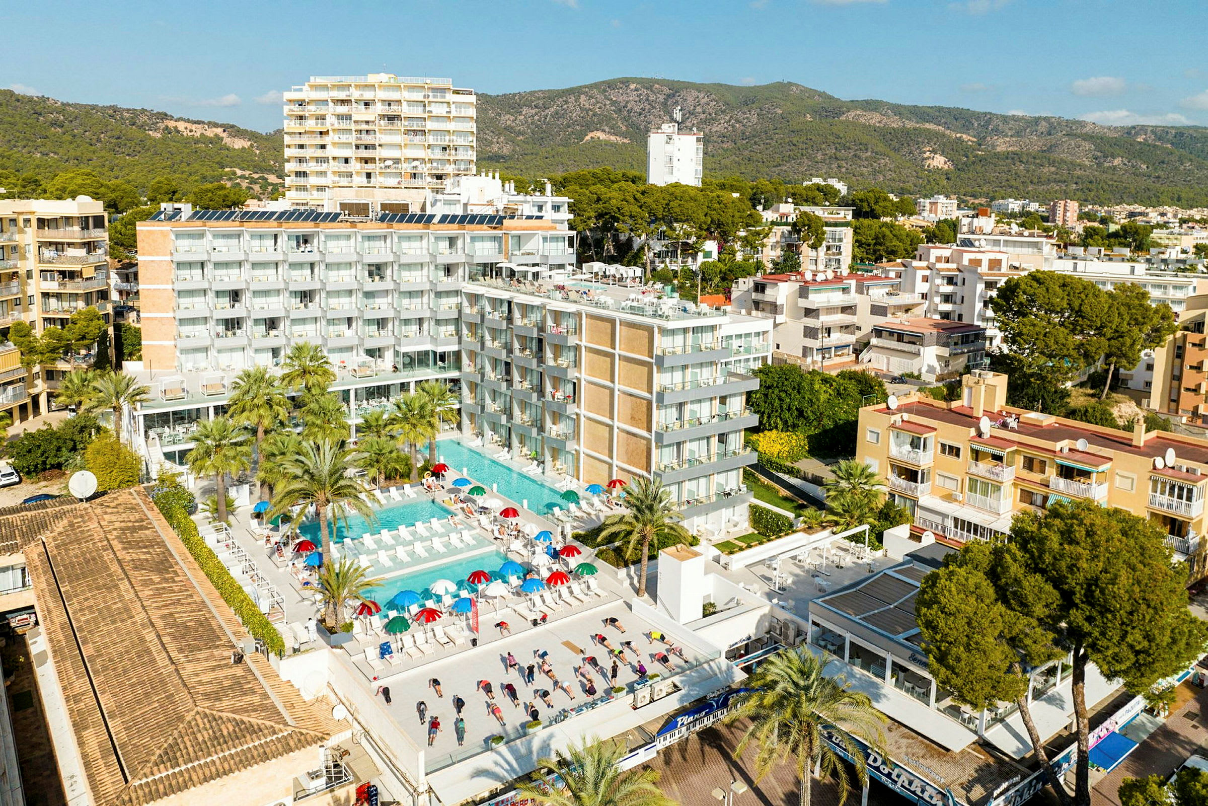Luftfoto af en hotelbygning med poolområde og liggestole. Det omkringliggende landskab med grønt og flere bygninger ses i baggrunden. (AI-genereret tekst)