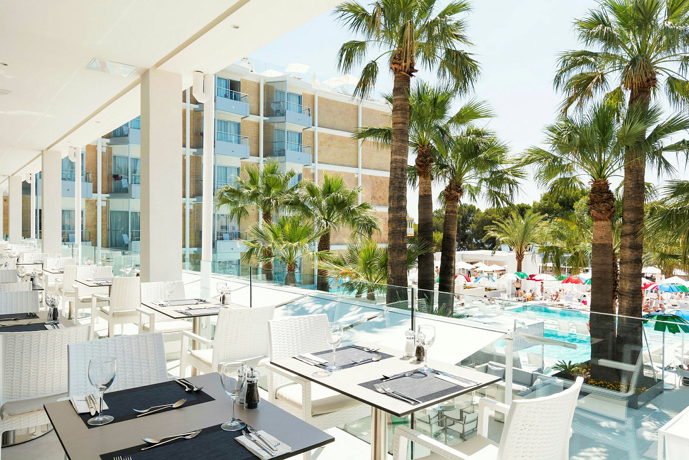 En terrasse med udendørs servering, borde og stole er placeret nær glasrækværk med udsigt over en pool og palmer. I baggrunden ses en bygning med altaner. (AI-genereret tekst)