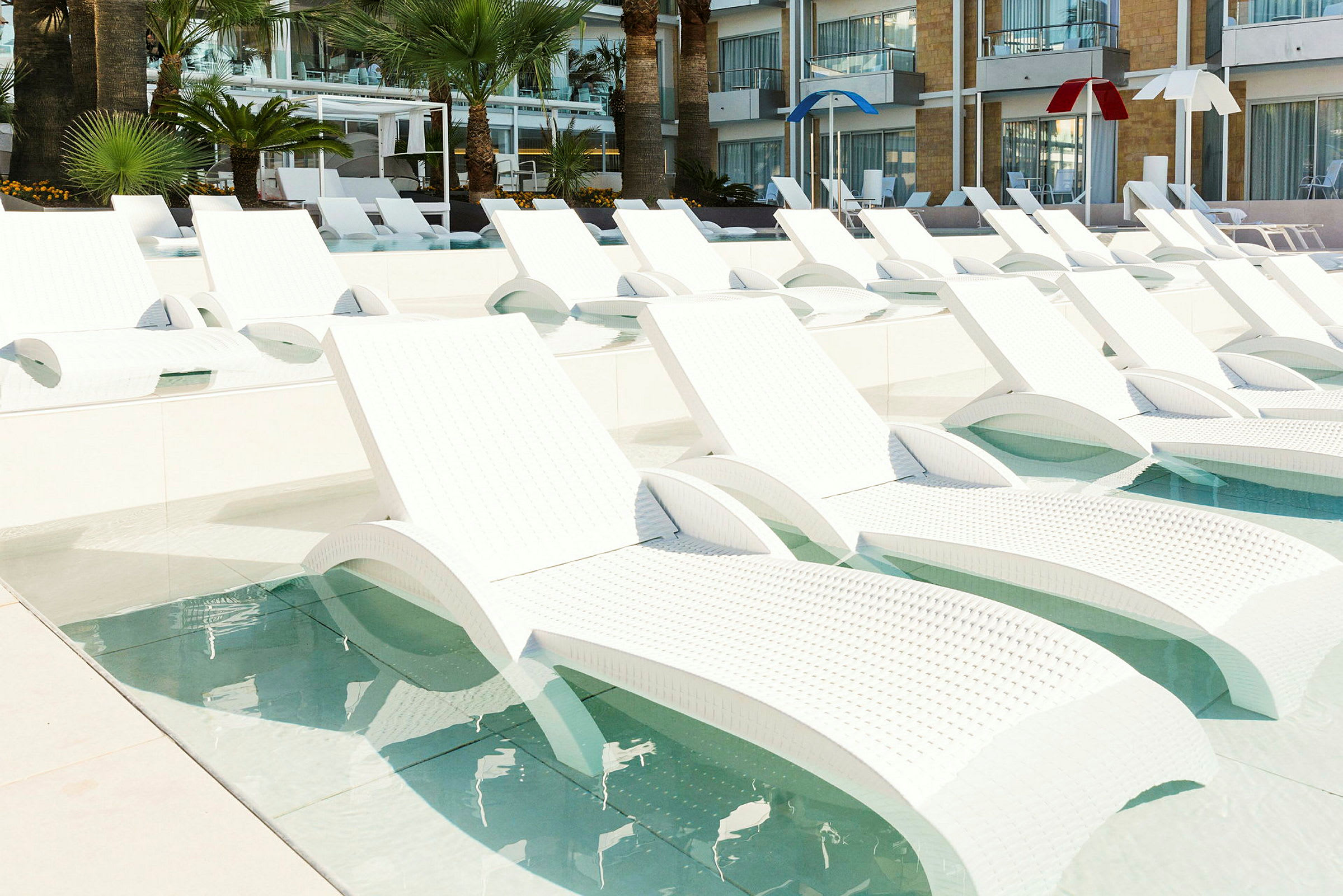 Billede af flere hvide liggestole placeret i en lav pool, med palmer og hotelbygning i baggrunden. (AI-genereret tekst)