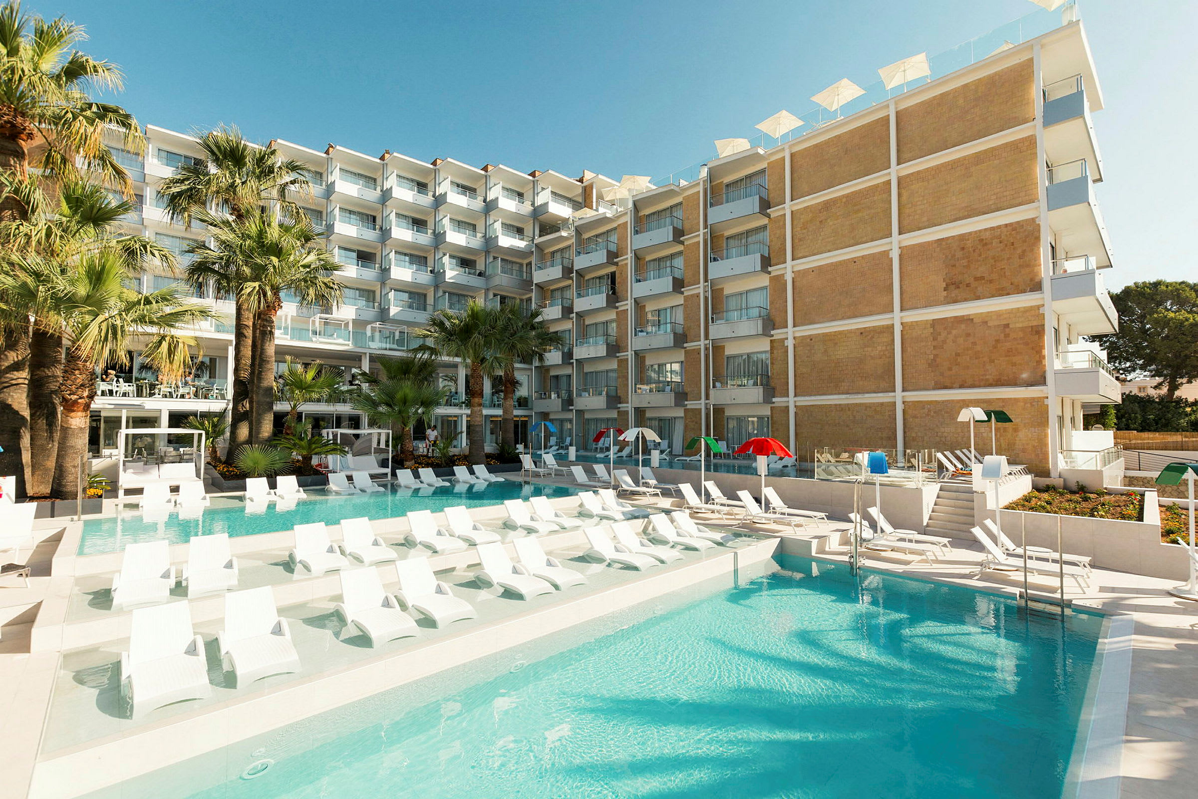 Et hotel med en pool omgivet af liggestole og grønne palmer. (AI-genereret tekst)