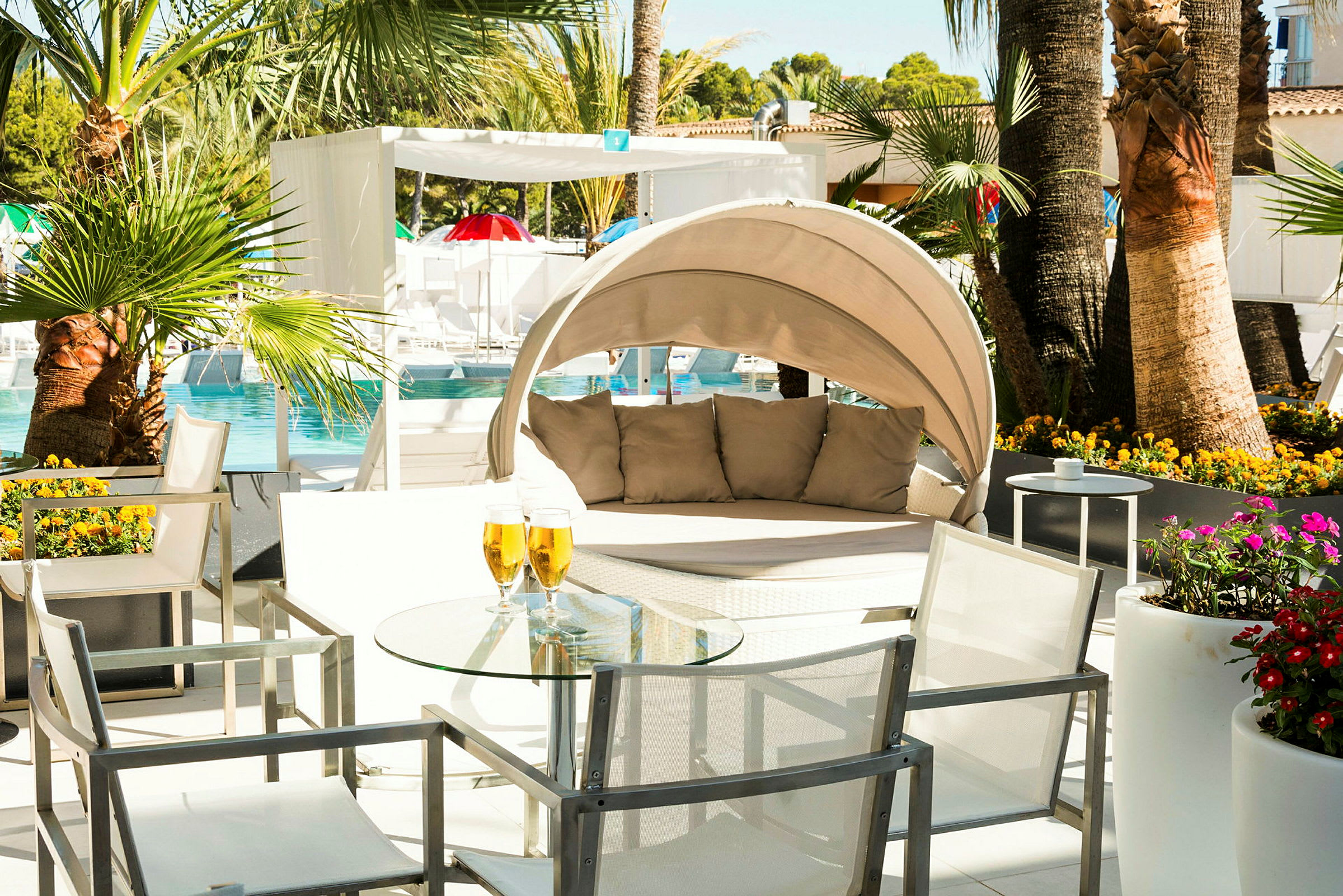 En solrig terrasse med en pool i baggrunden. Der er hvide møbler, blomster og en rund solseng med puder. (AI-genereret tekst)