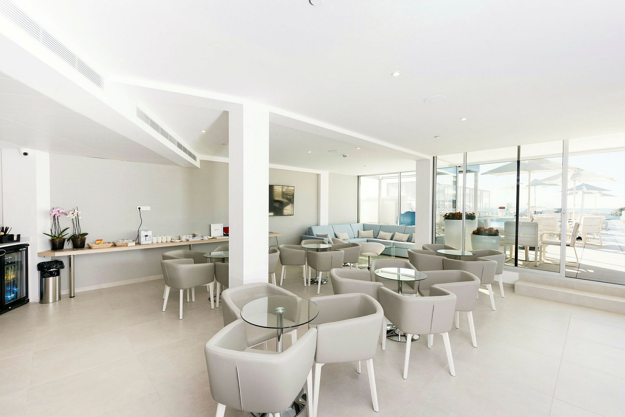 En moderne lounge med grå møbler og glasborde. En serveringsområde med drikkevarer og snacks er synlig til venstre, og store glasvinduer åbner op til en udendørsterrasse. (AI-genereret tekst)