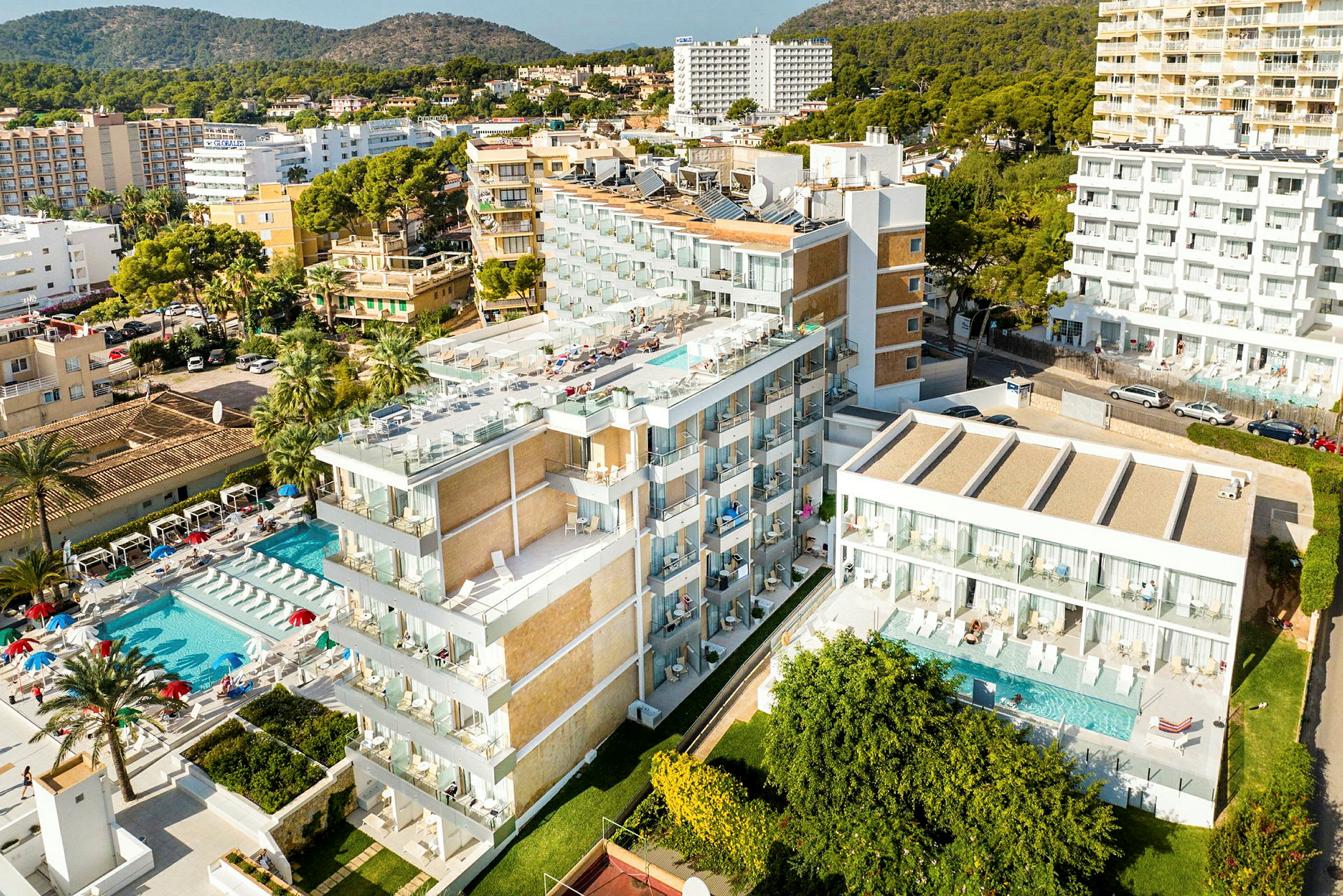 Billede af et hotelkompleks med poolområde og omgivende bygninger, omgivet af grønne områder og ujævnt terræn. (AI-genereret tekst)