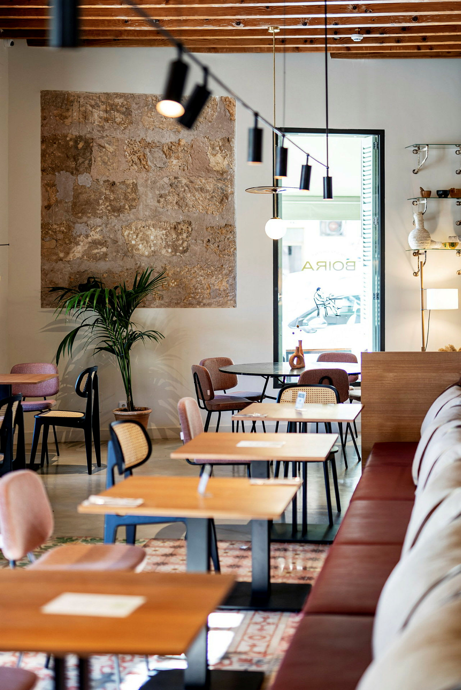 En moderne café med træborde og stole samt en plante ved en væg med sten. Lyset fra vinduet oplyser rummet. (AI-genereret tekst)