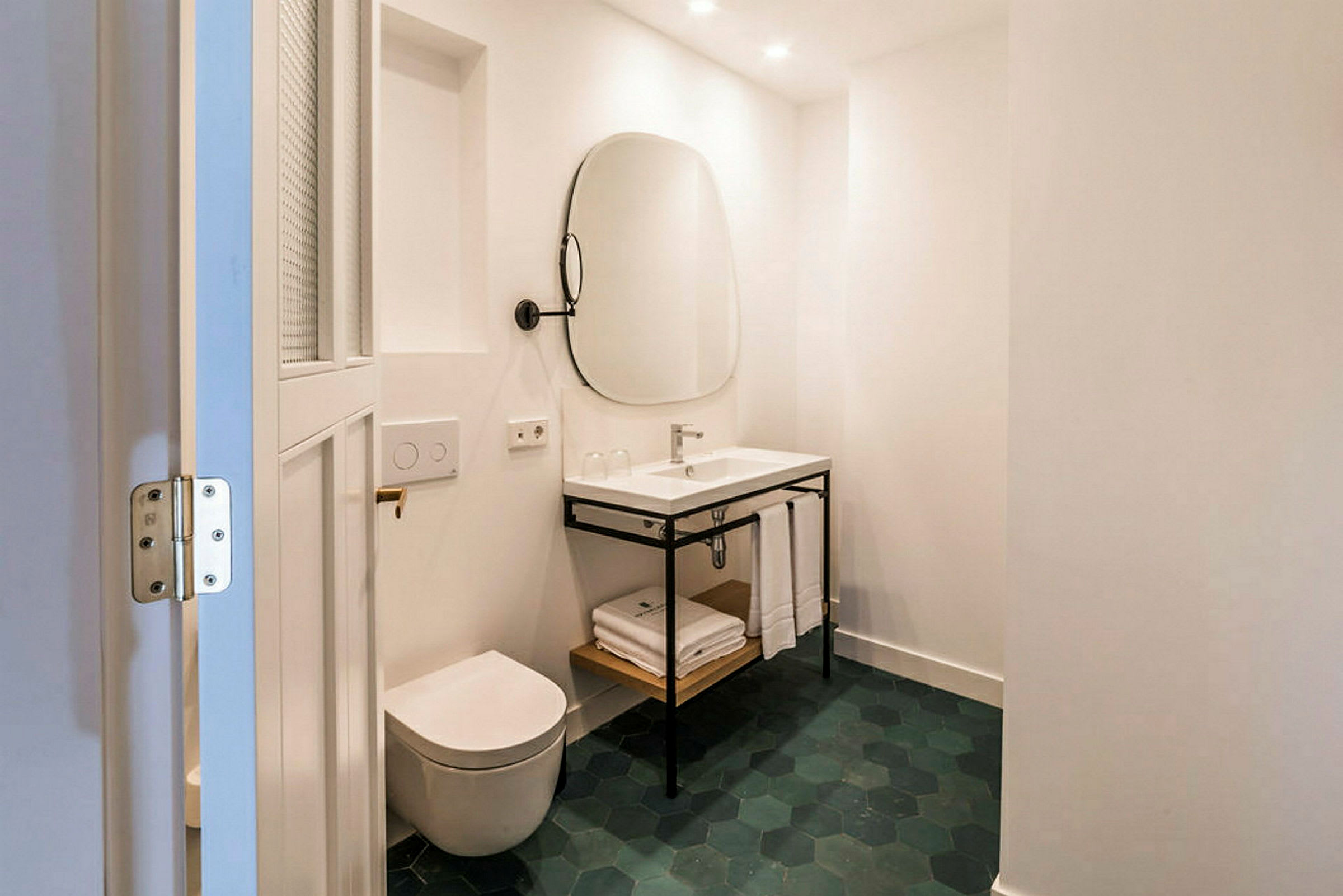 Et moderne badeværelse med et hvidt toilet og en håndvask med tilhørende spejl. Gulvet har sekskantede grønne fliser. (AI-genereret tekst)