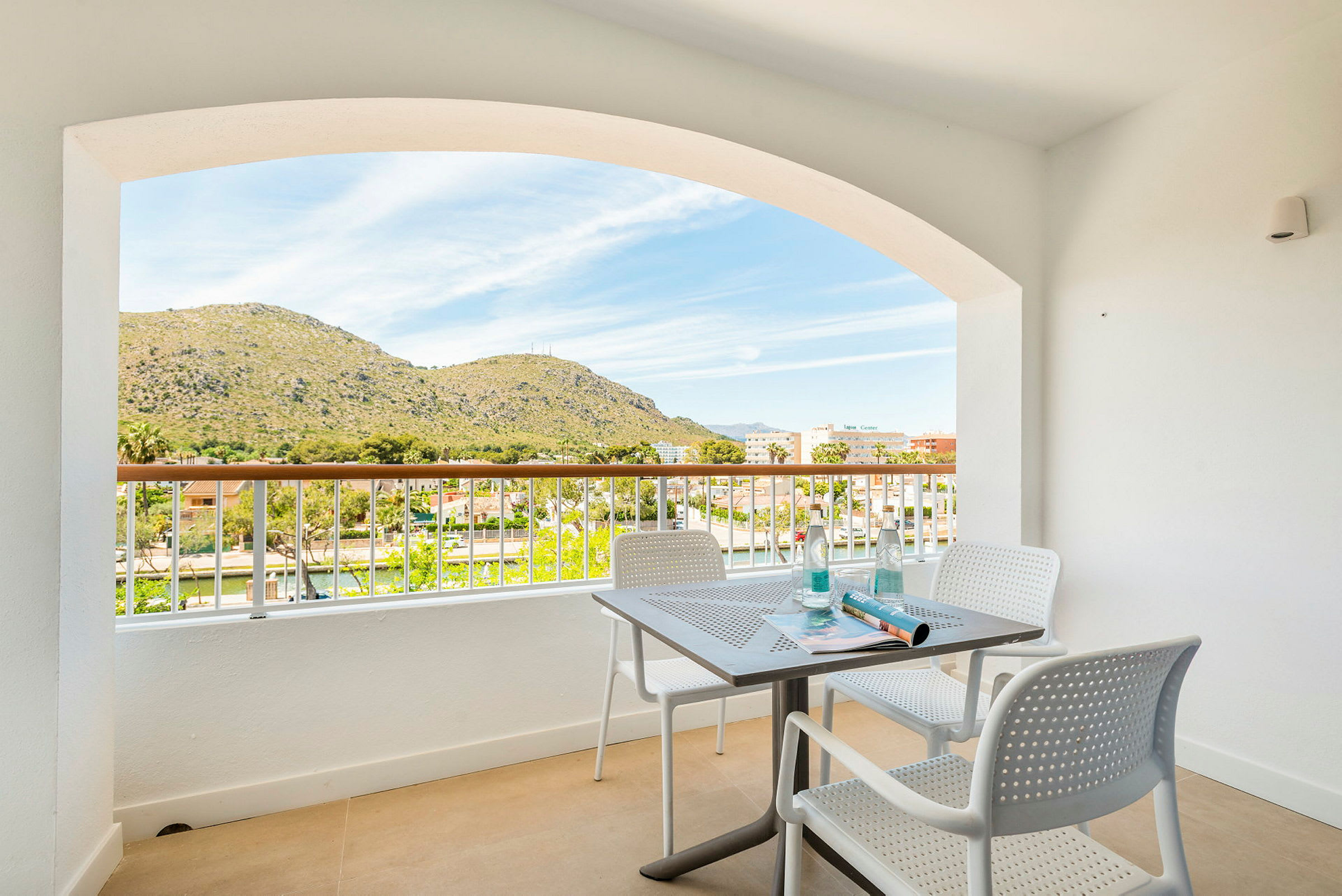 Udsigt fra en balkon med bord og stole mod et bjerglandskab. (AI-genereret tekst)