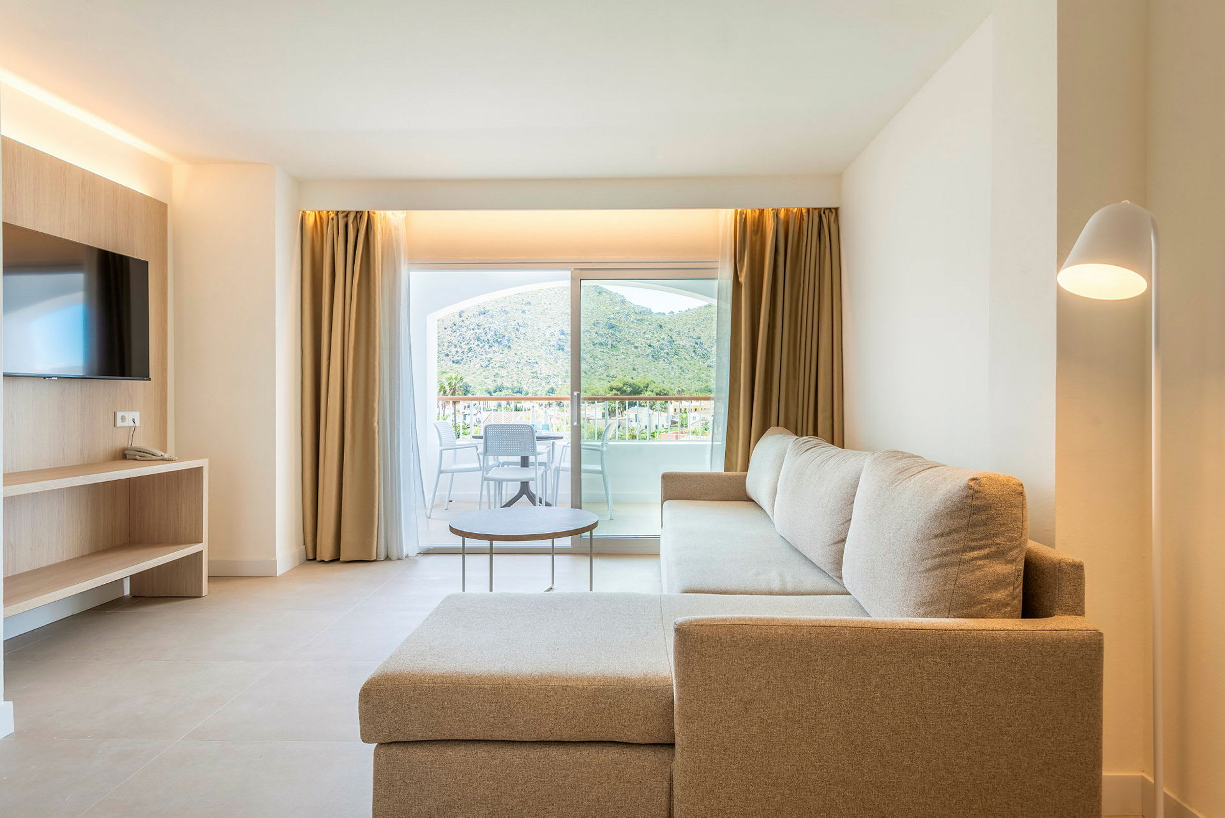 Stueinteriør med beige sofa, TV og glasdør med udsigt til en terrasse og bjerge. (AI-genereret tekst)