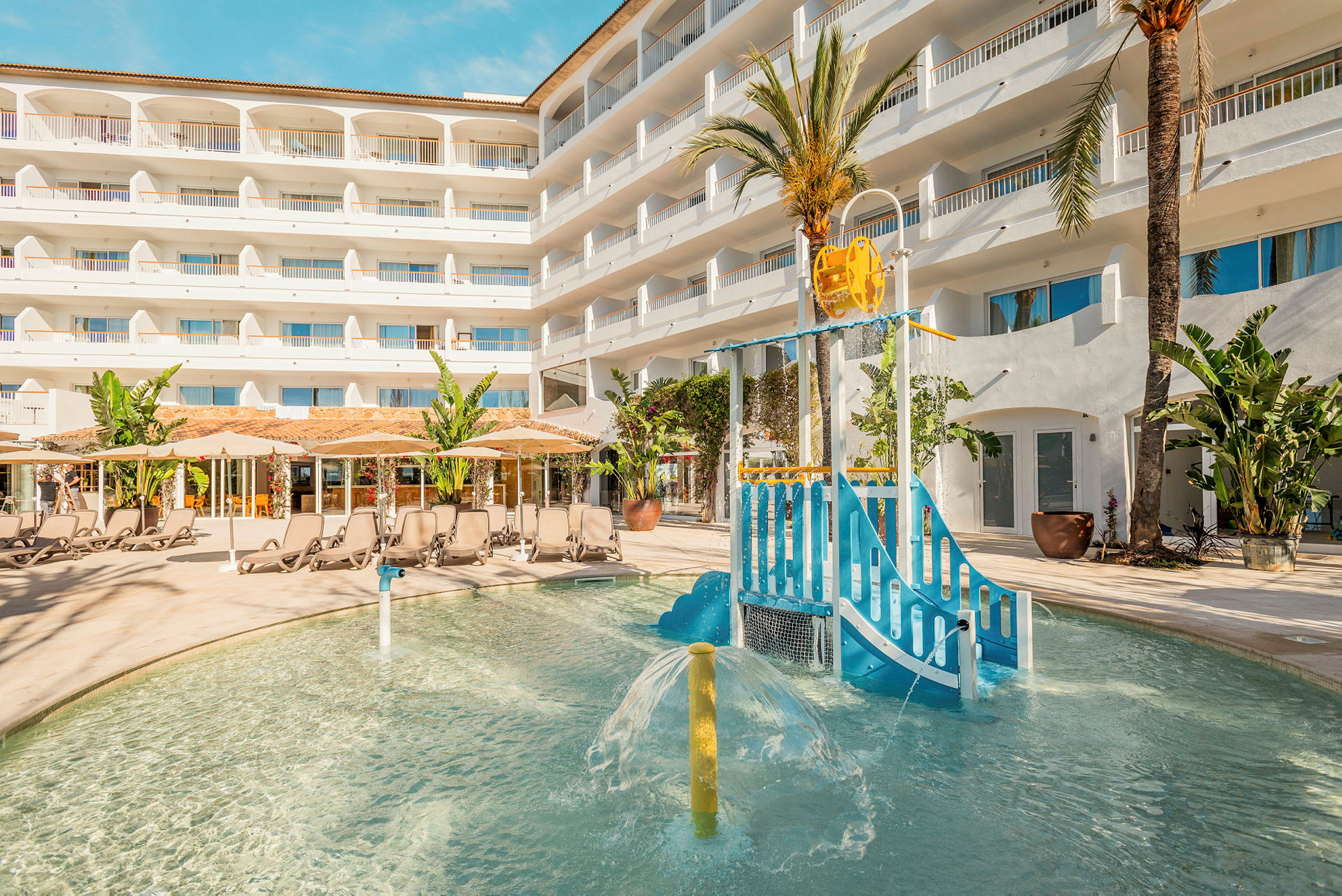 Udendørs poolområde med legeplads for børn og liggestole foran hotelbygning. (AI-genereret tekst)