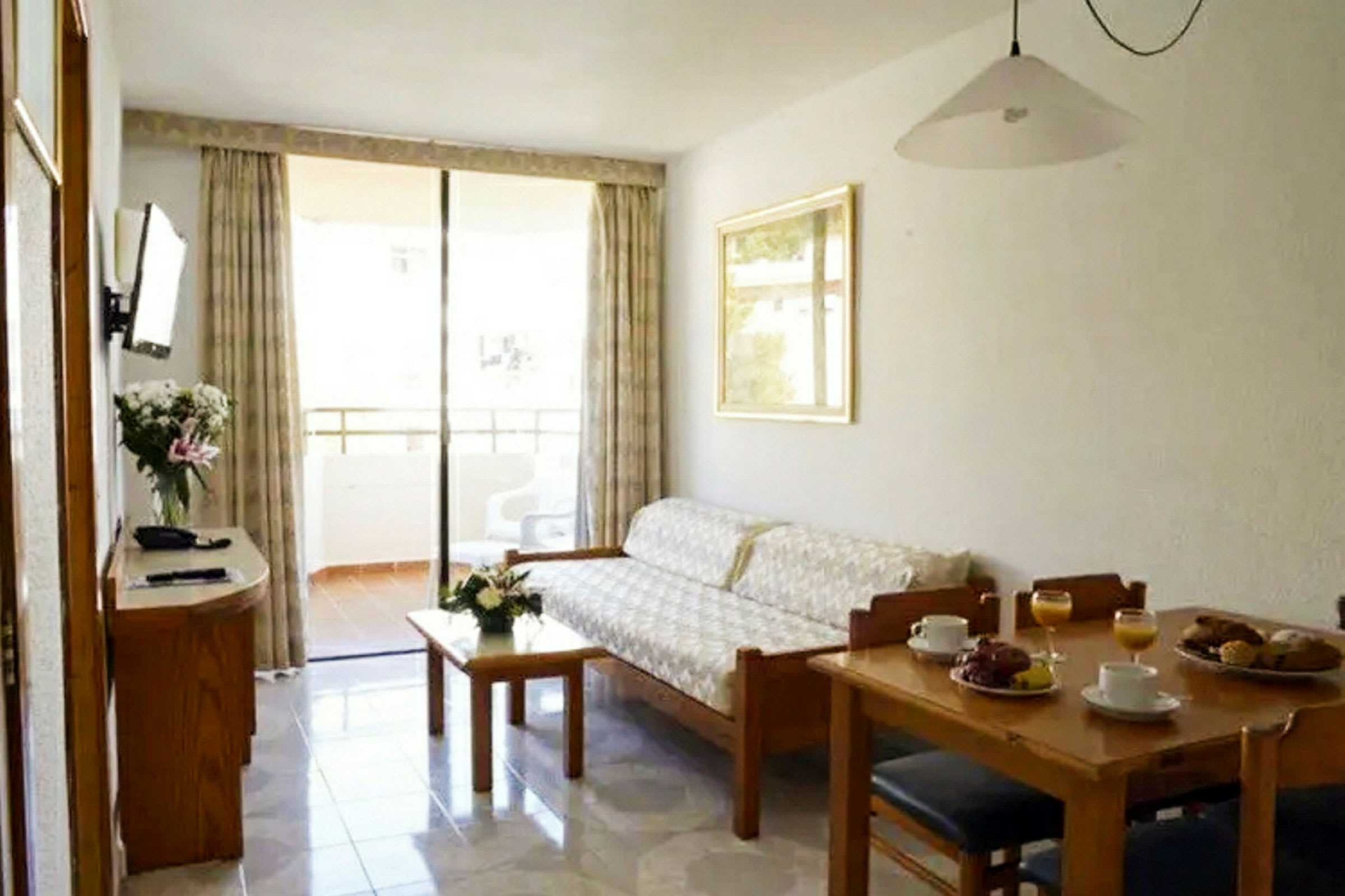 En lejlighed med en stue, der indeholder en sofa, et bord med blomster, en spiseplads med opdækket bord og en balkon i baggrunden. (AI-genereret tekst)