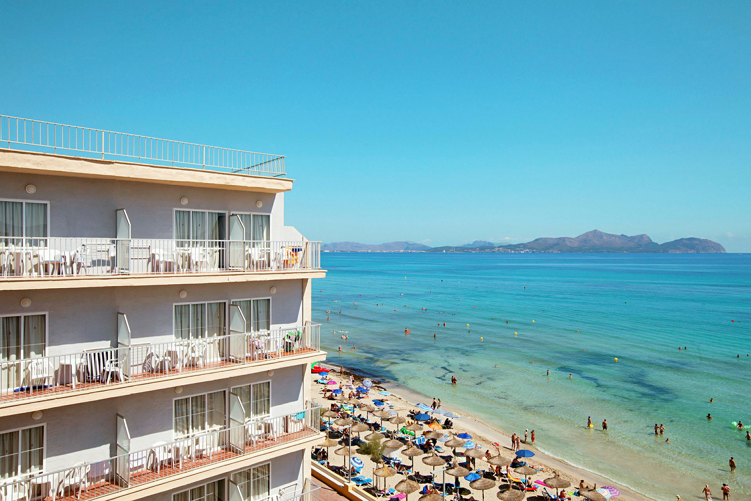 Et strandhotel nær en sandstrand med parasoller, og mennesker bader i klart blåt vand. (AI-genereret tekst)